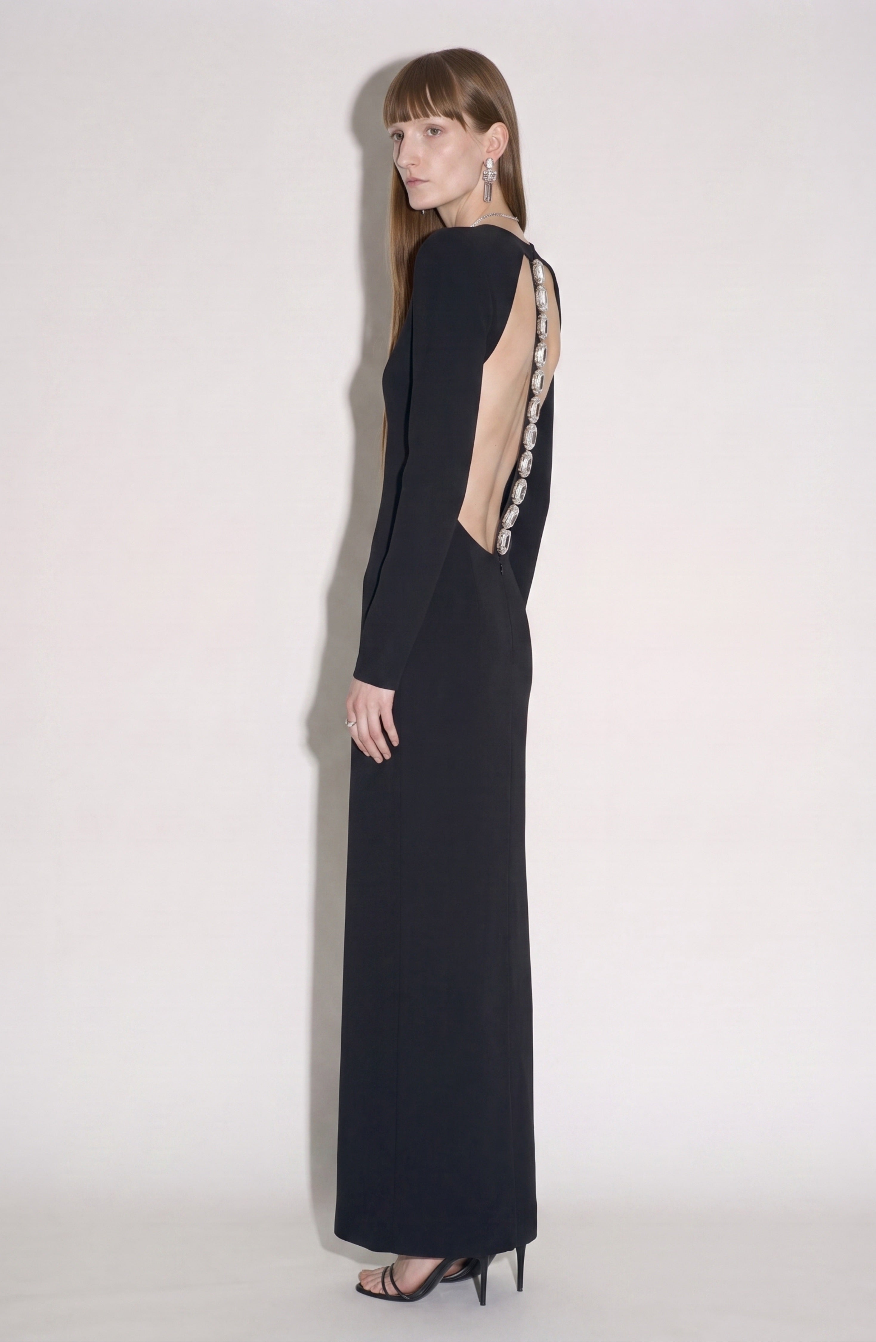 A woman wearing the LOULOU Back Crepe Maxi Dress—a long black crepe gown featuring a deep open back and decorative buttons—stands in profile against a plain light background, styled with black heels and statement earrings.