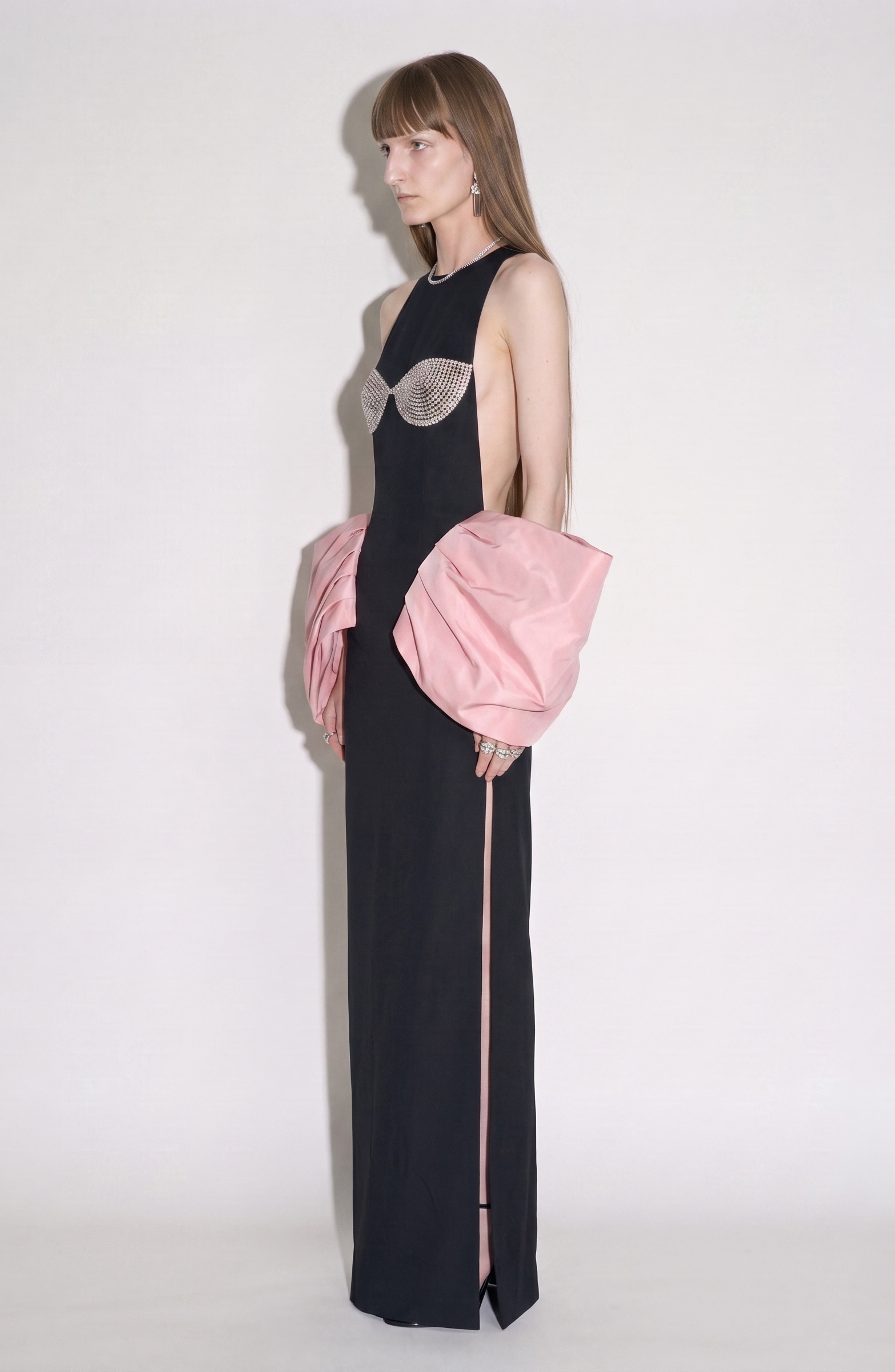 A person stands sideways, wearing the LOULOU Balloon Sleeve Gown—a striking black column dress with a crystal-embellished bustier and dramatic pale pink balloon sleeves that drape elegantly at the sides. The background is plain white.