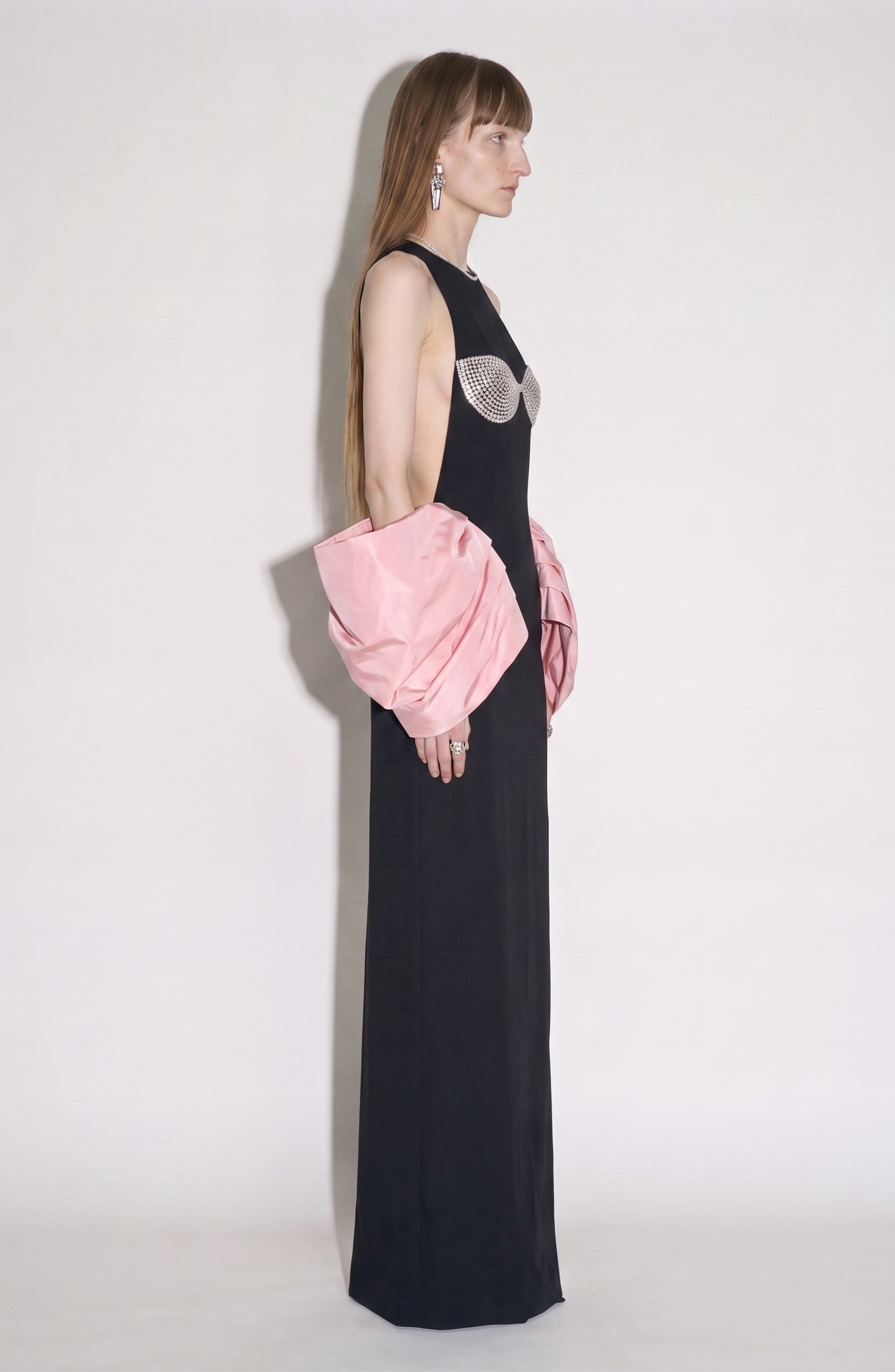 A person in the LOULOU Balloon Sleeve Gown, featuring a long black column silhouette with a crystal-embellished bustier and dramatic balloon sleeves, stands in profile against a plain light background.