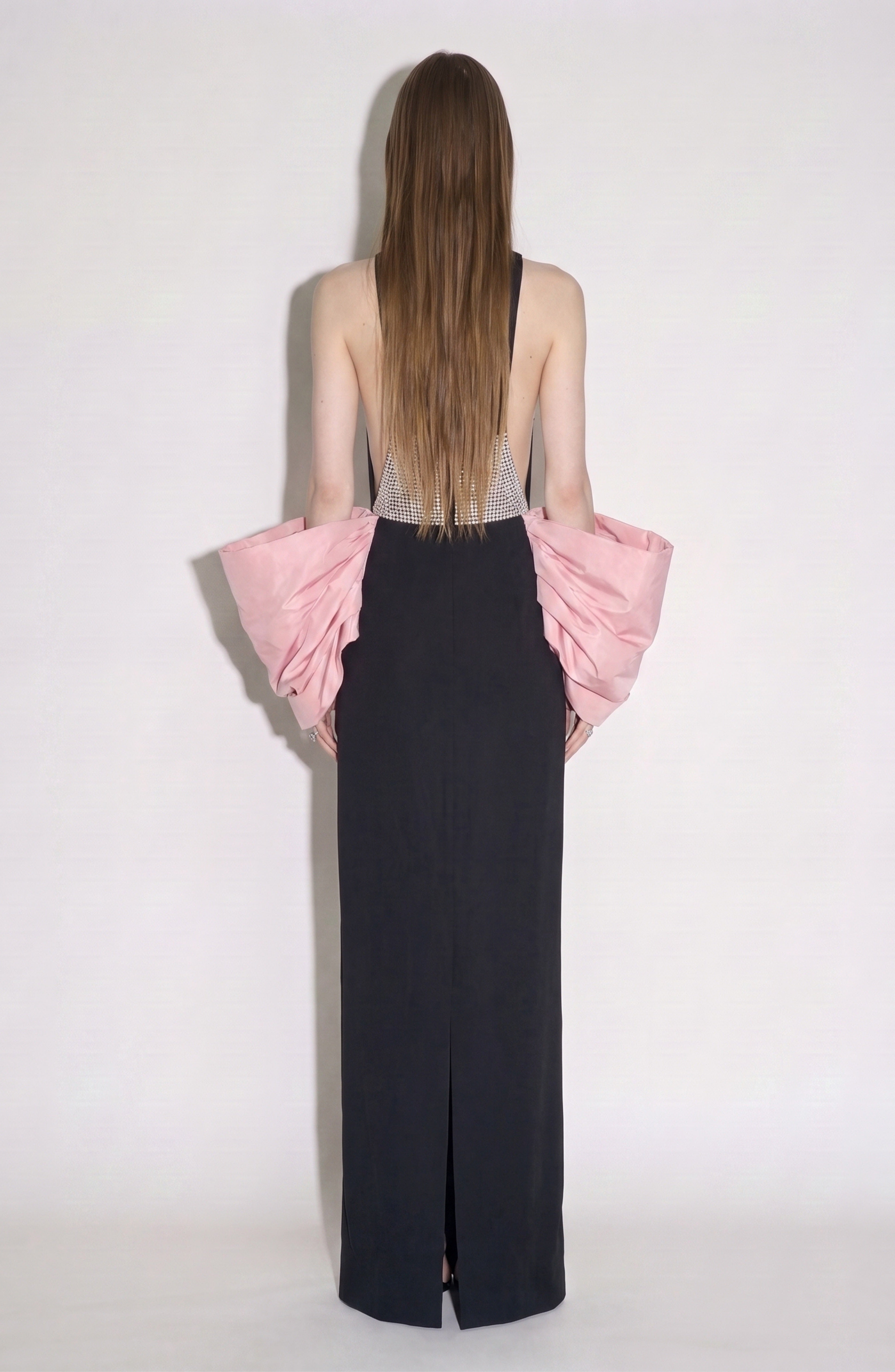 A person with long hair stands facing away, wearing the LOULOU Balloon Sleeve Gown—a black column dress featuring a low back and dramatic ruffled pink balloon sleeves. The background is plain and light-colored.