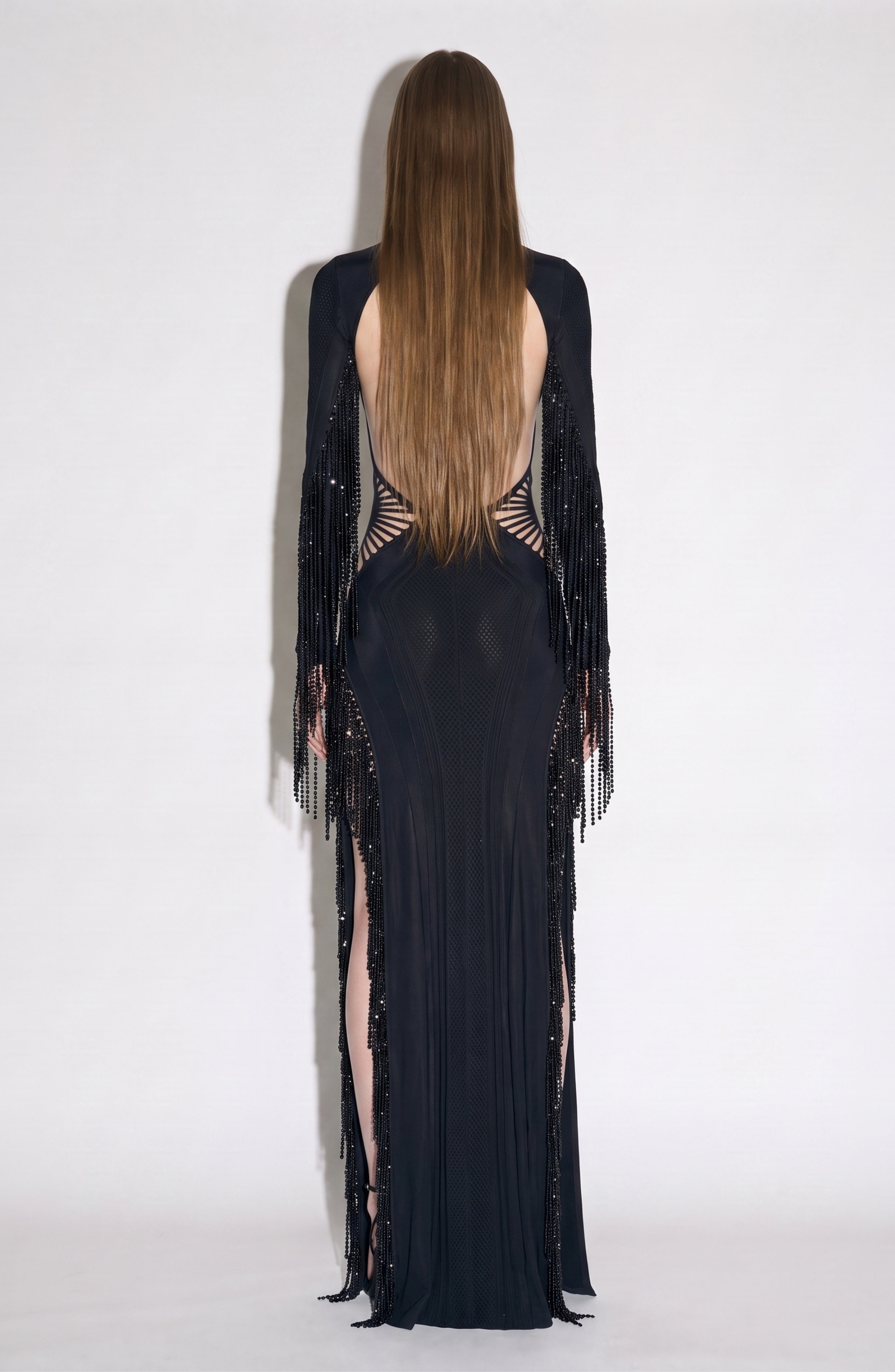 A person with long straight hair stands facing away against a plain light background, wearing the LOULOU Beaded Fringe Cut-Out Gown—a jet black maxi dress featuring an open back, beaded fringe sleeves, and bold geometric paneling and cutouts.