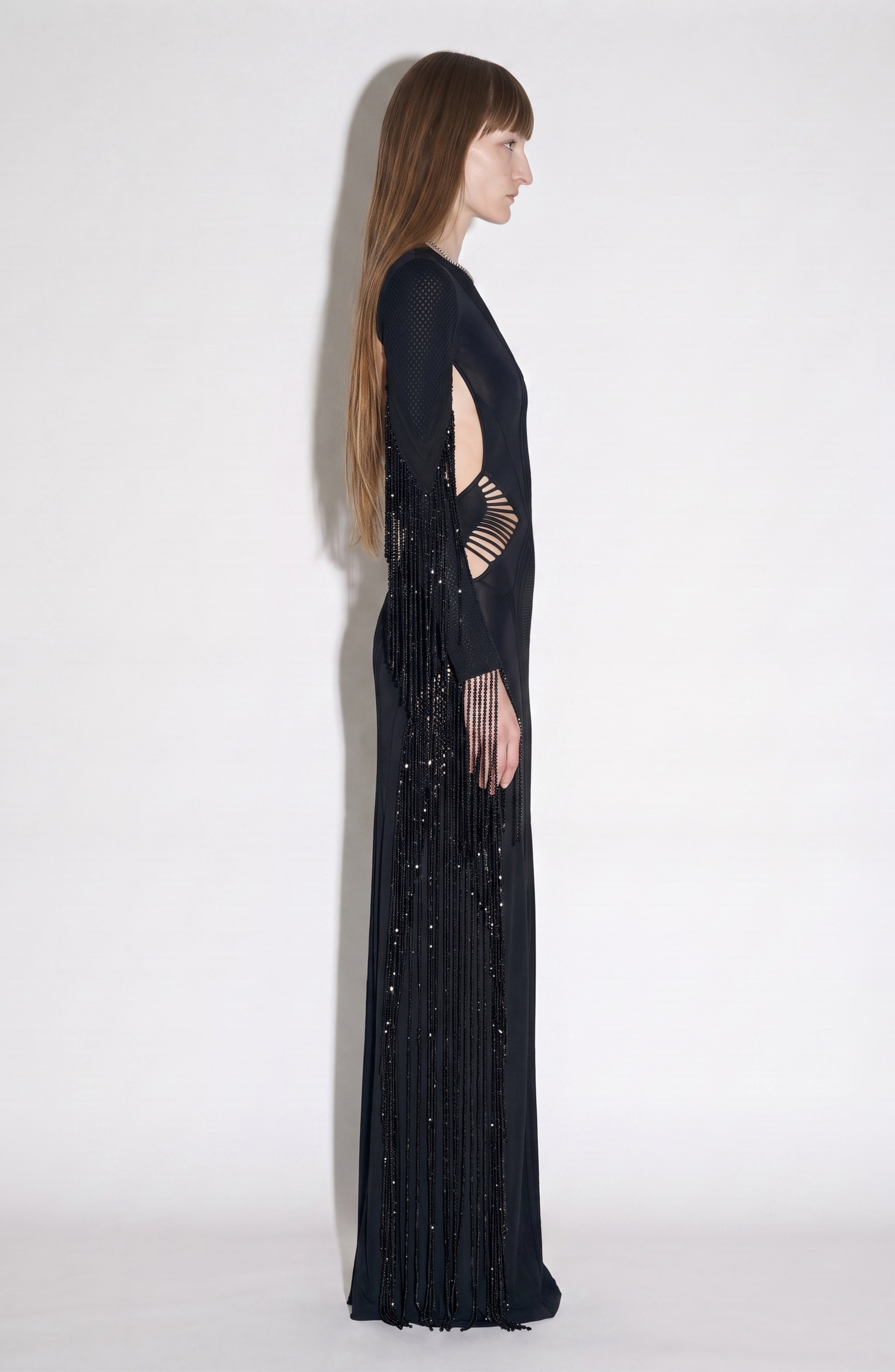 A woman stands in profile against a plain light background, wearing the LOULOU Beaded Fringe Cutout Maxi Dress—an elegant long black dress featuring geometric cutouts, beaded fringe, and intricate bead detailing. Her straight brown hair is worn loose as she looks ahead.