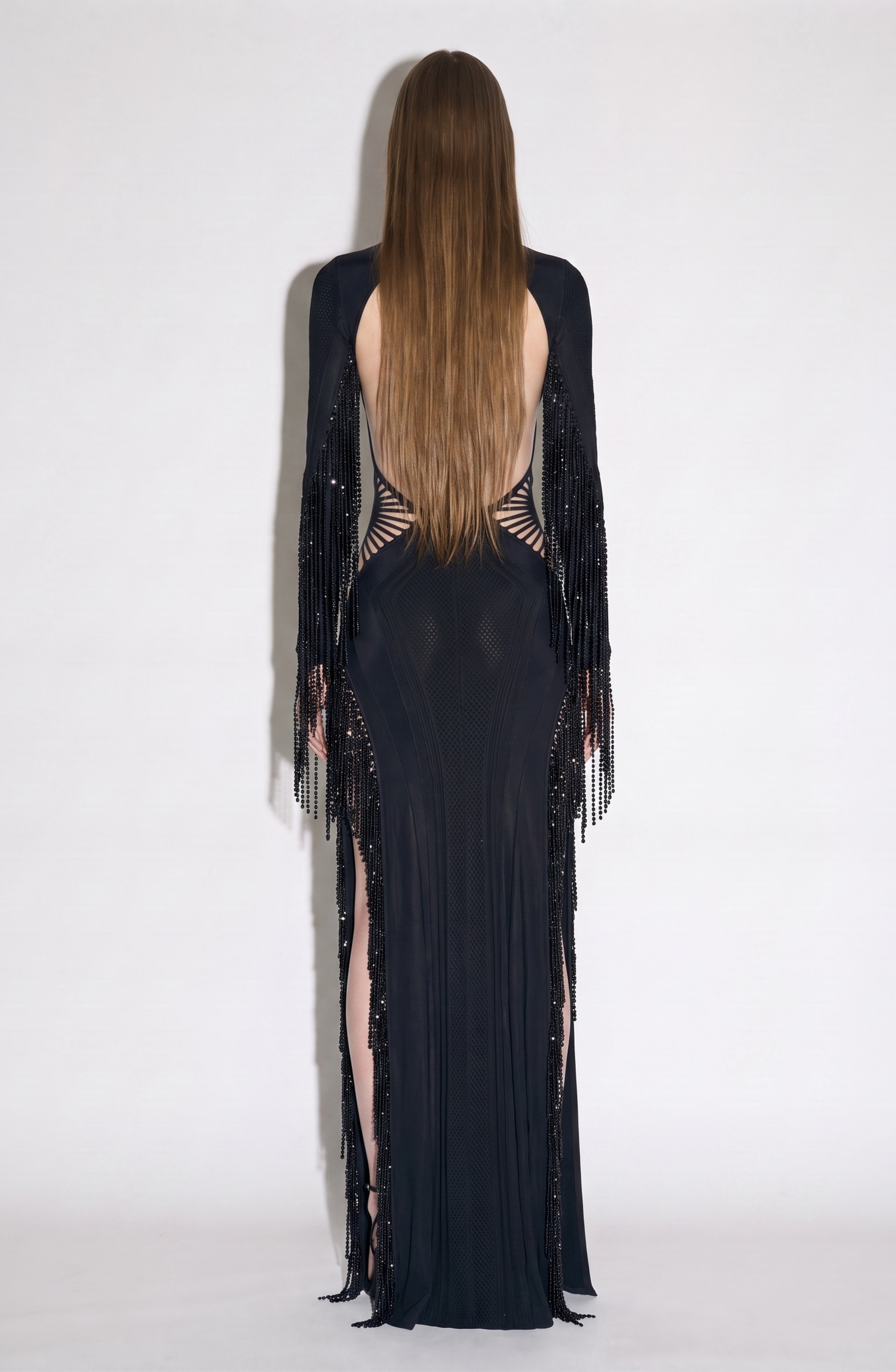 A person with long straight hair stands facing away, wearing the LOULOU Beaded Fringe Cutout Maxi Dress—featuring beaded fringe details on the arms, an open back, and subtle geometric cutouts—set against a plain light background.