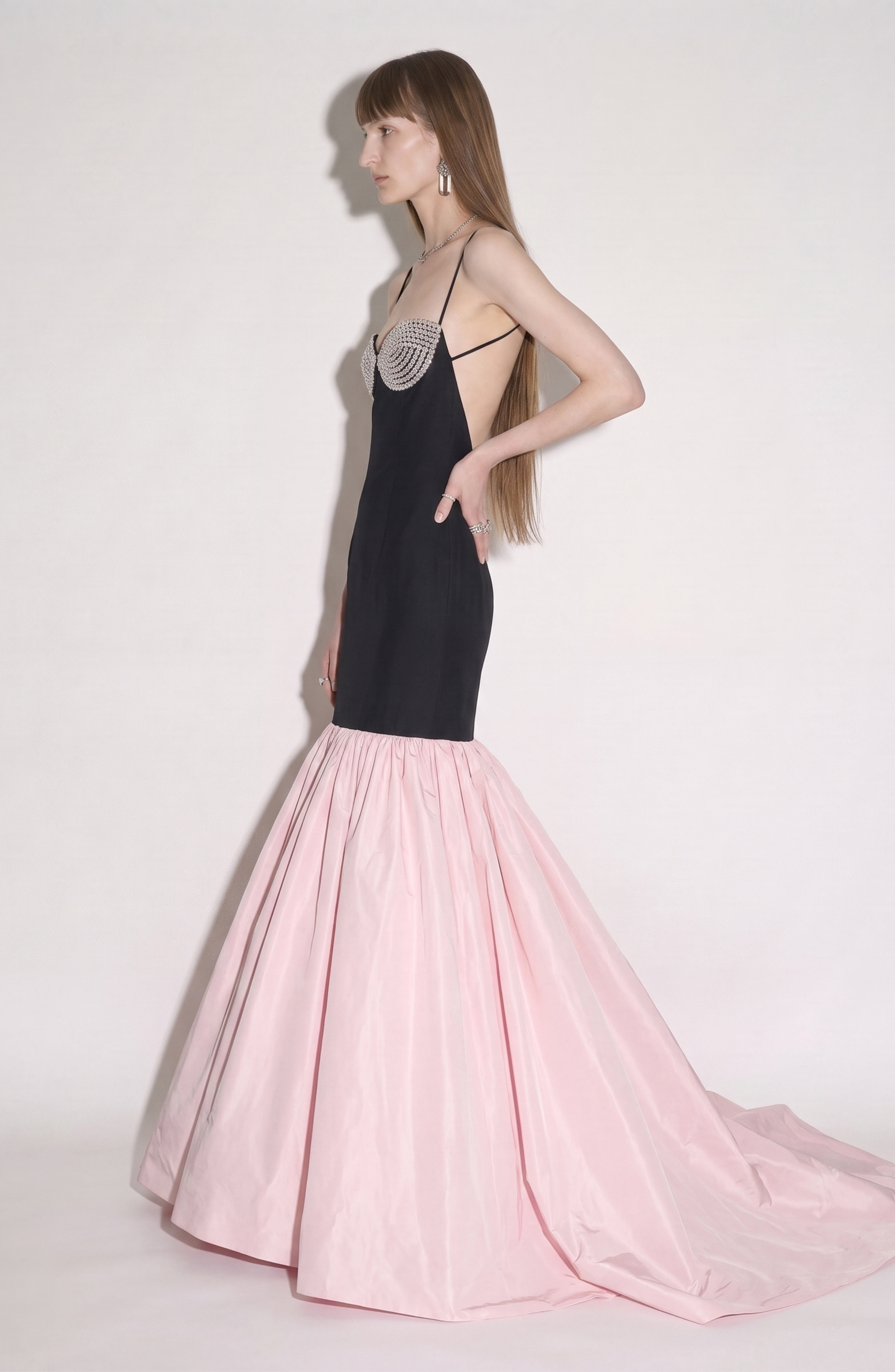 A woman stands in profile against a plain light background, wearing the LOULOU Contrast Mermaid Gown. The dress features a fitted black bodice with thin straps and a crystal-embellished bustier, flowing into a voluminous pink mermaid skirt with a dramatic floor-sweeping train.