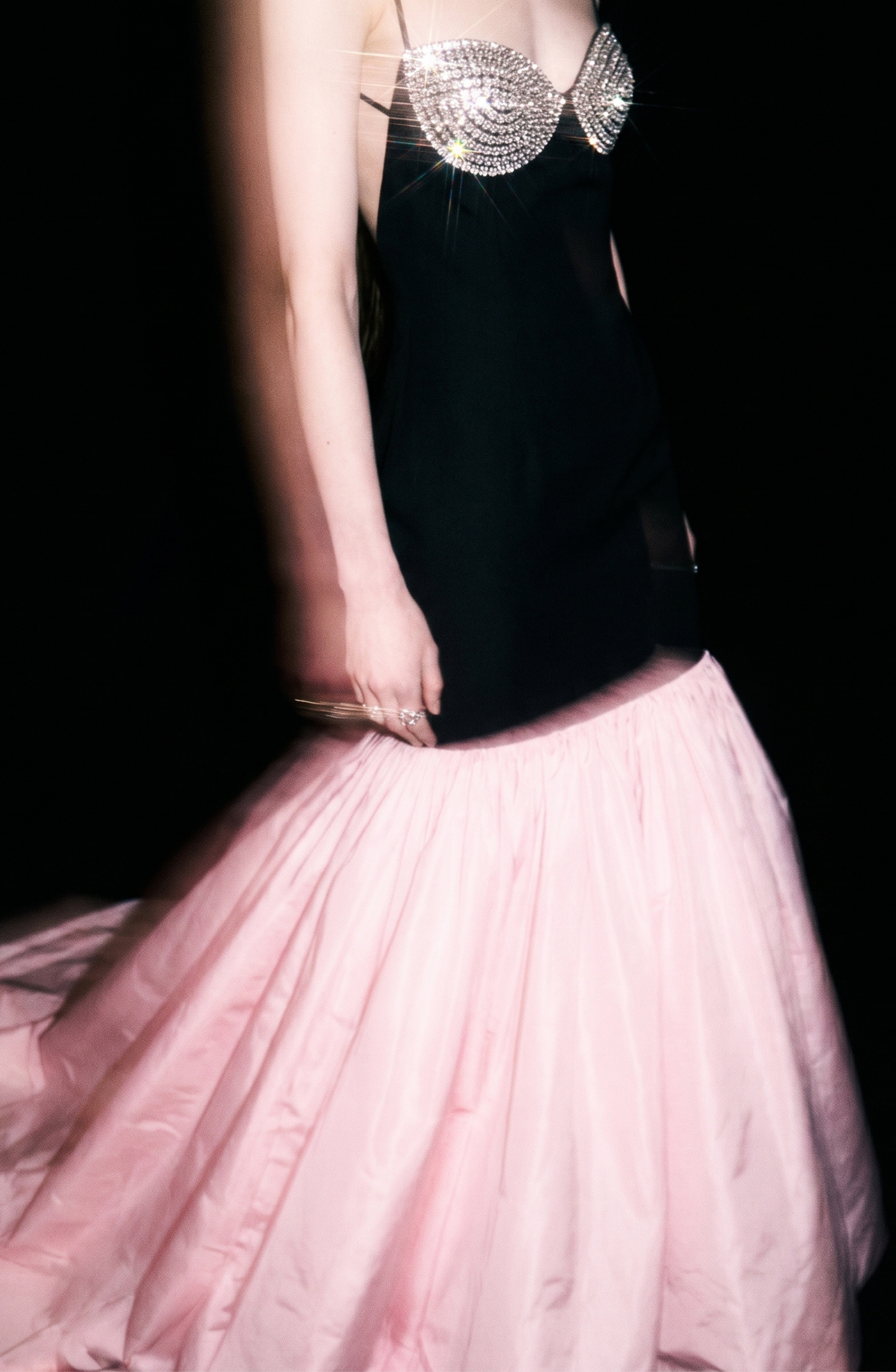 A person wearing the LOULOU Contrast Mermaid Gown—a strapless black evening gown with a crystal-embellished bustier and a dramatic, voluminous pink skirt featuring a floor-sweeping train—stands against a dark background, with motion blur on the arms.
