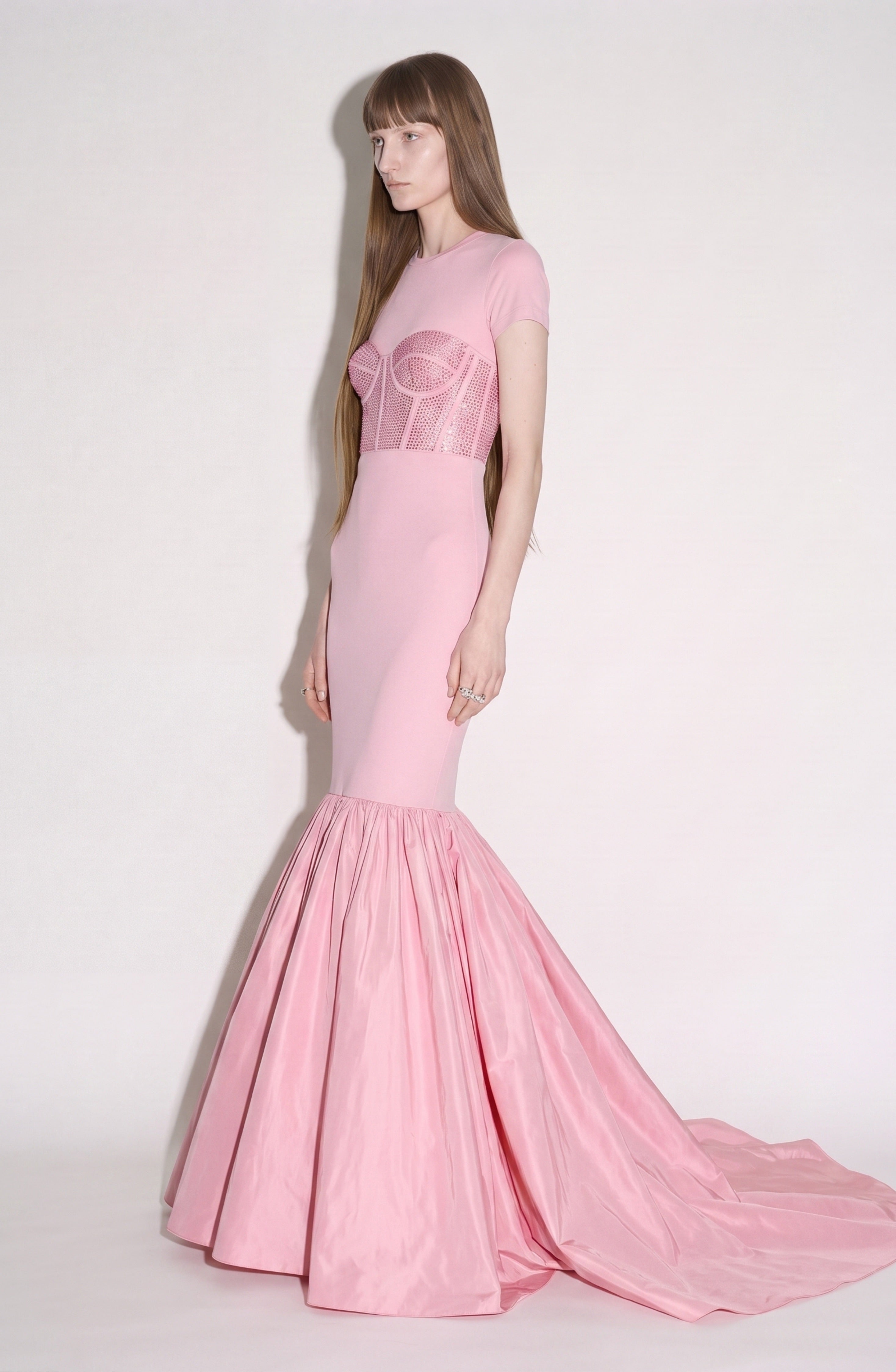 A person with long straight hair wears the LOULOU Corset Mermaid Gown, a fitted, floor-length pink dress featuring a crystal-embellished corset and a flared, voluminous taffeta skirt, standing against a plain light background.