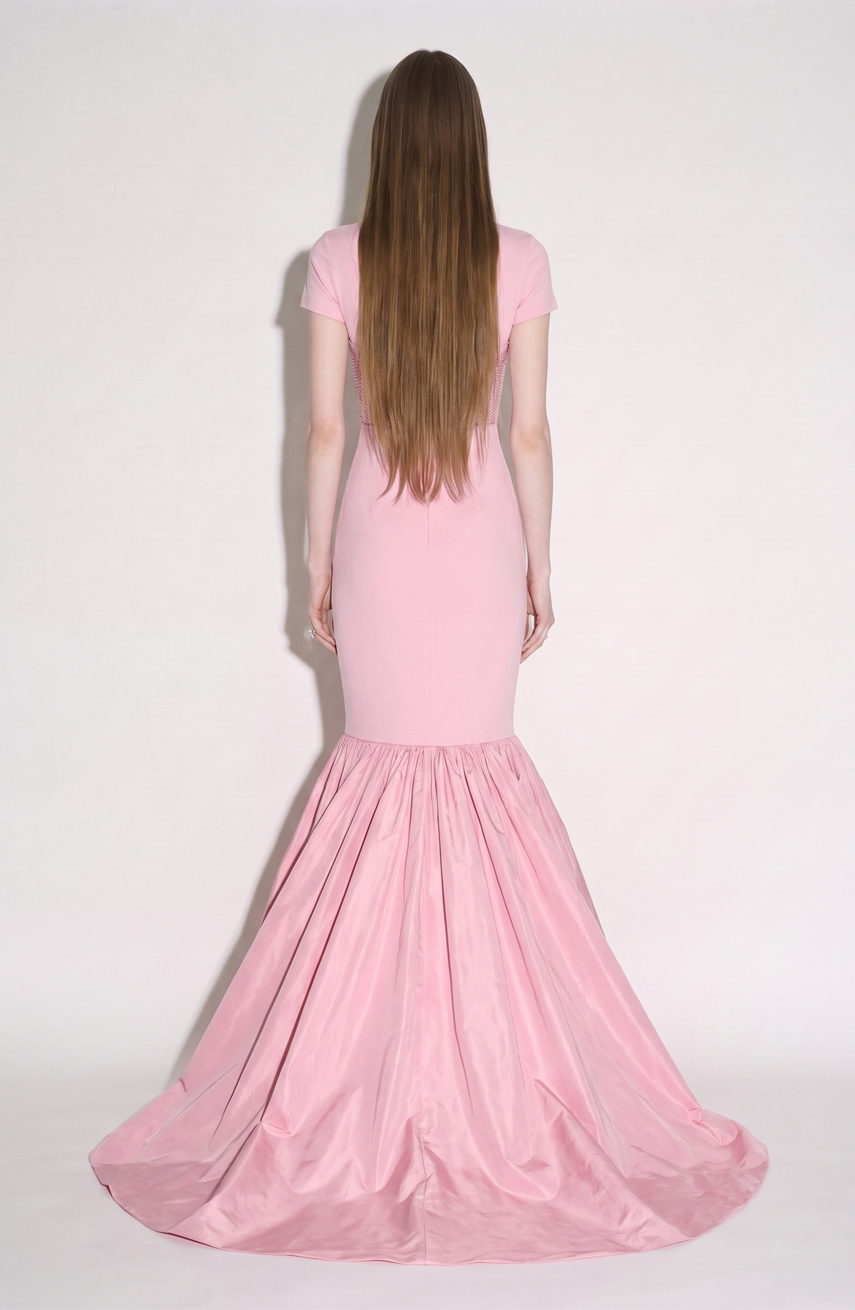 A person with long brown hair stands facing away, wearing the LOULOU Corset Mermaid Gown—a fitted light pink dress featuring a crystal-embellished corset and a flared, gathered taffeta skirt that extends to the floor—set against a plain background.