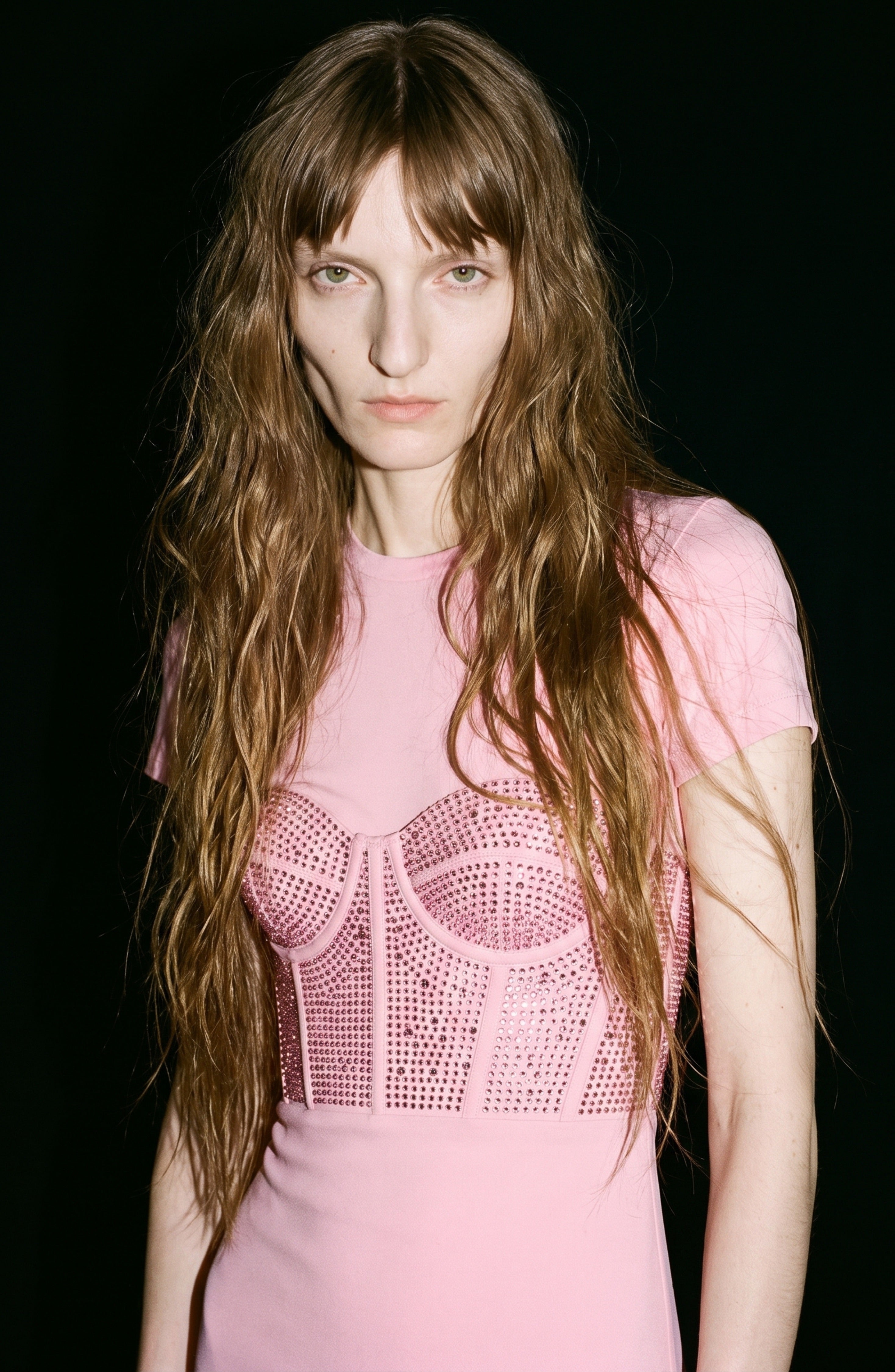 A person with long wavy brown hair wears the LOULOU Corset Mermaid Gown, a light pink dress adorned with studs and crystal-embellished corset detailing, while standing against a black background.