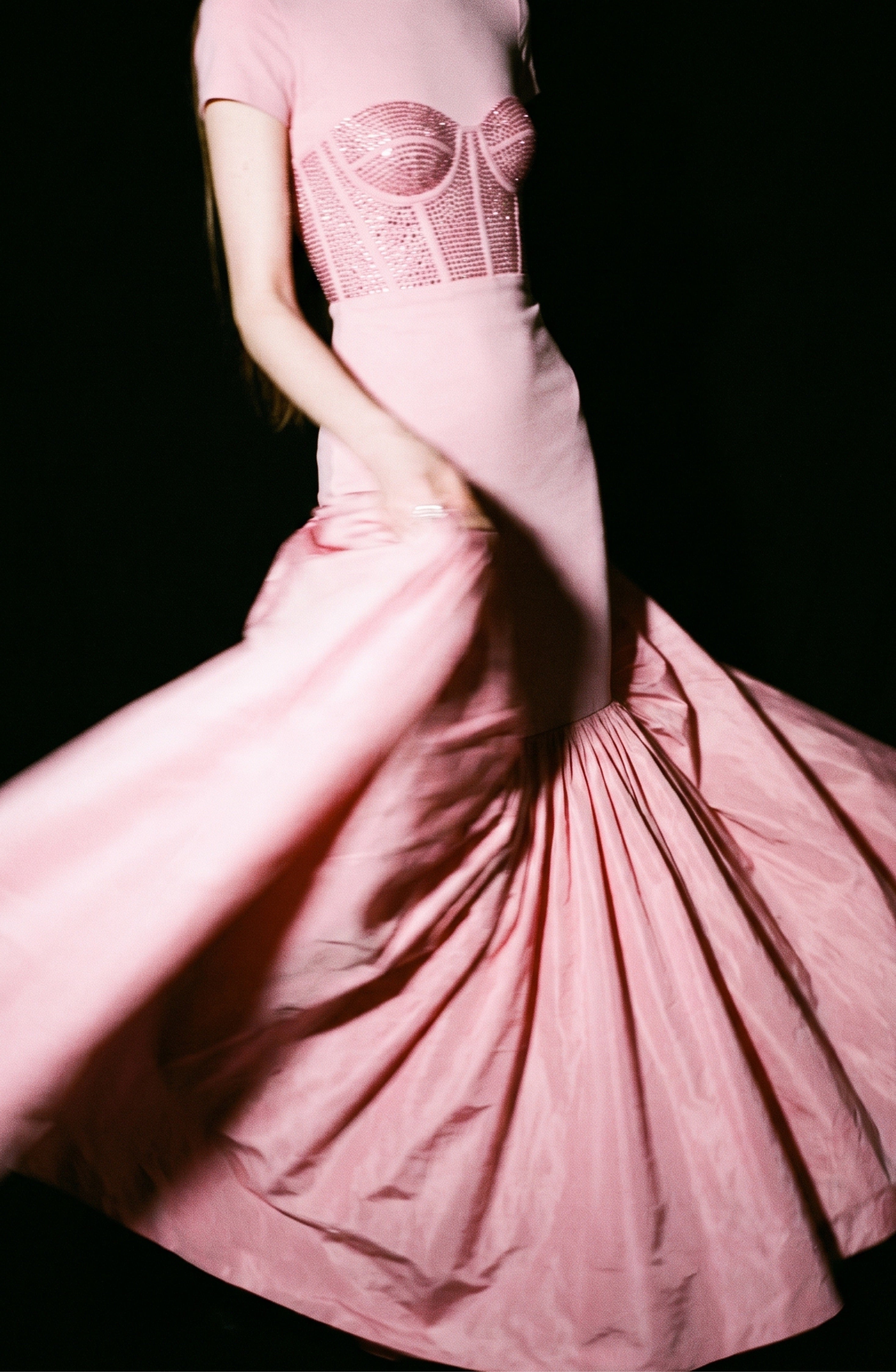 A woman wearing the LOULOU Corset Mermaid Gown, featuring a fitted, crystal-embellished corset and a flared taffeta skirt, twirls gracefully as the skirt fans out. Against a black background, the pink gown stands out brilliantly.