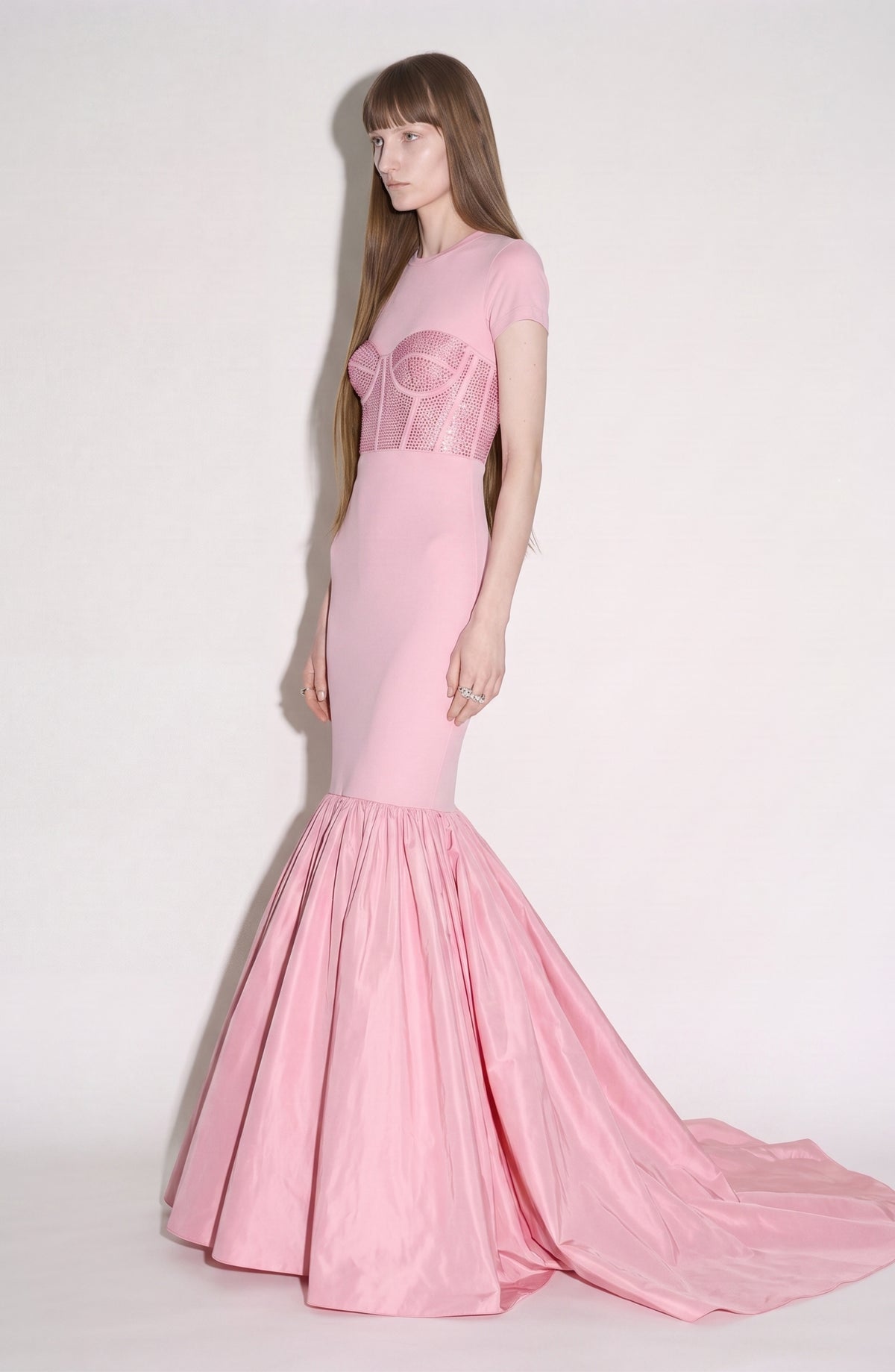 A woman stands sideways against a plain, light background, wearing the LOULOU Corset Mermaid Gown—a fitted mermaid silhouette crafted from pale pink silk taffeta with a structured bodice and flared skirt.