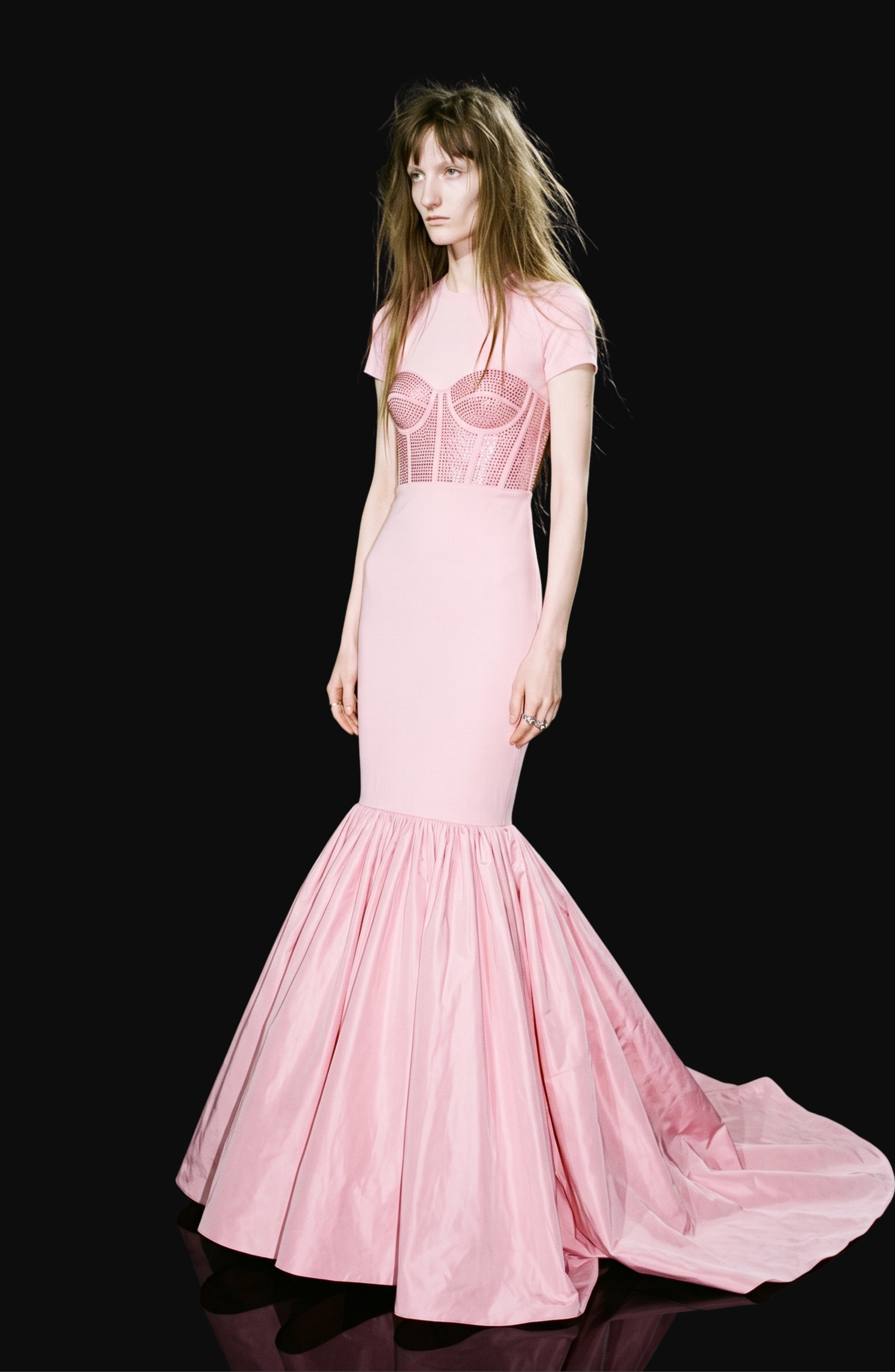 A woman stands against a black background, wearing the LOULOU Corset Mermaid Gown—a pale pink, short-sleeve, fitted gown with a crystal-embellished corset-style bodice and a structured silhouette that flares into a floor-length taffeta mermaid skirt.