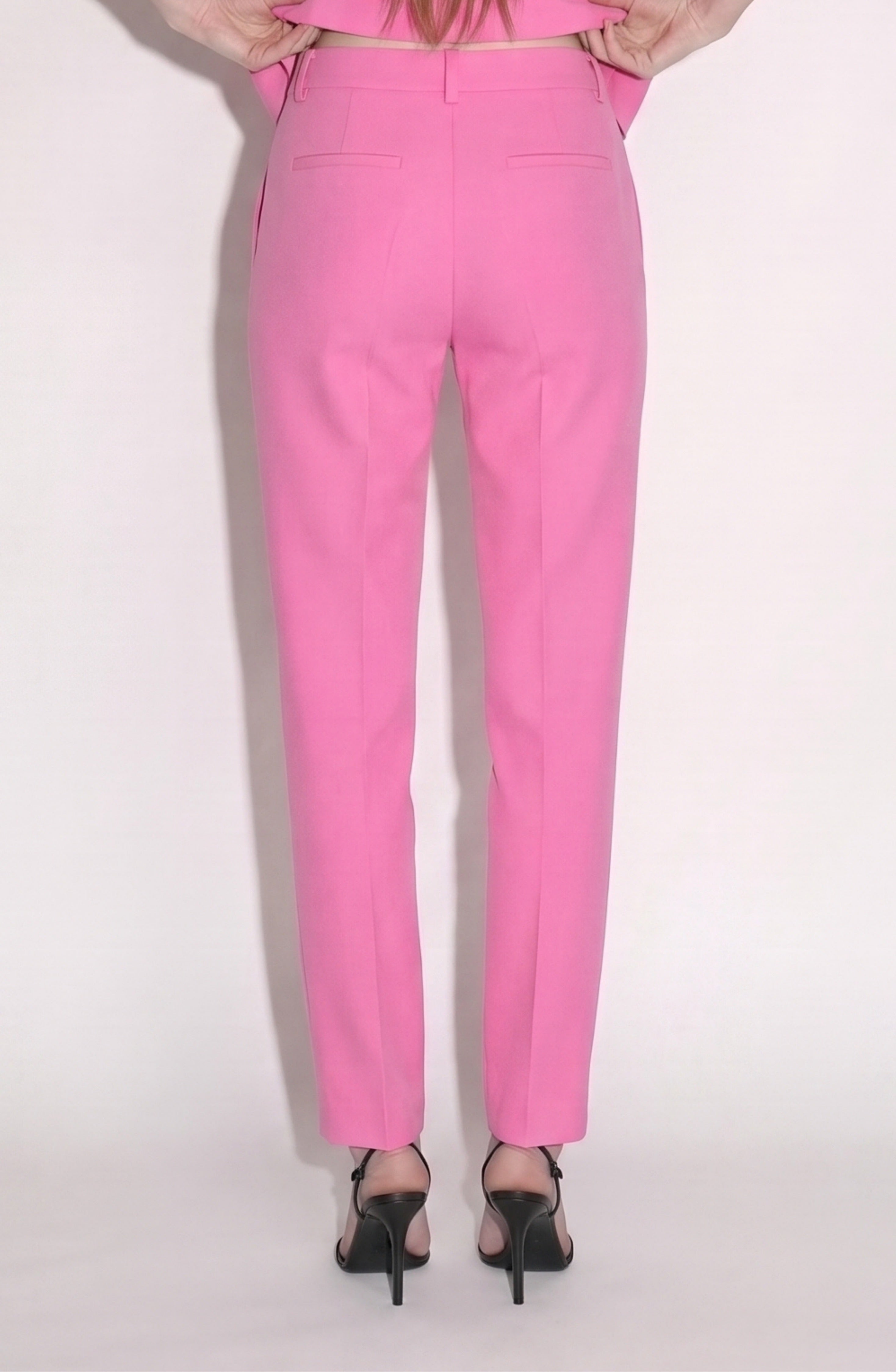 A person stands against a light background, shown from the waist down in a cropped silhouette, wearing LOULOU's bubblegum pink Cropped Tailored Trousers and black high-heeled sandals, with their hands tucked into their pockets.