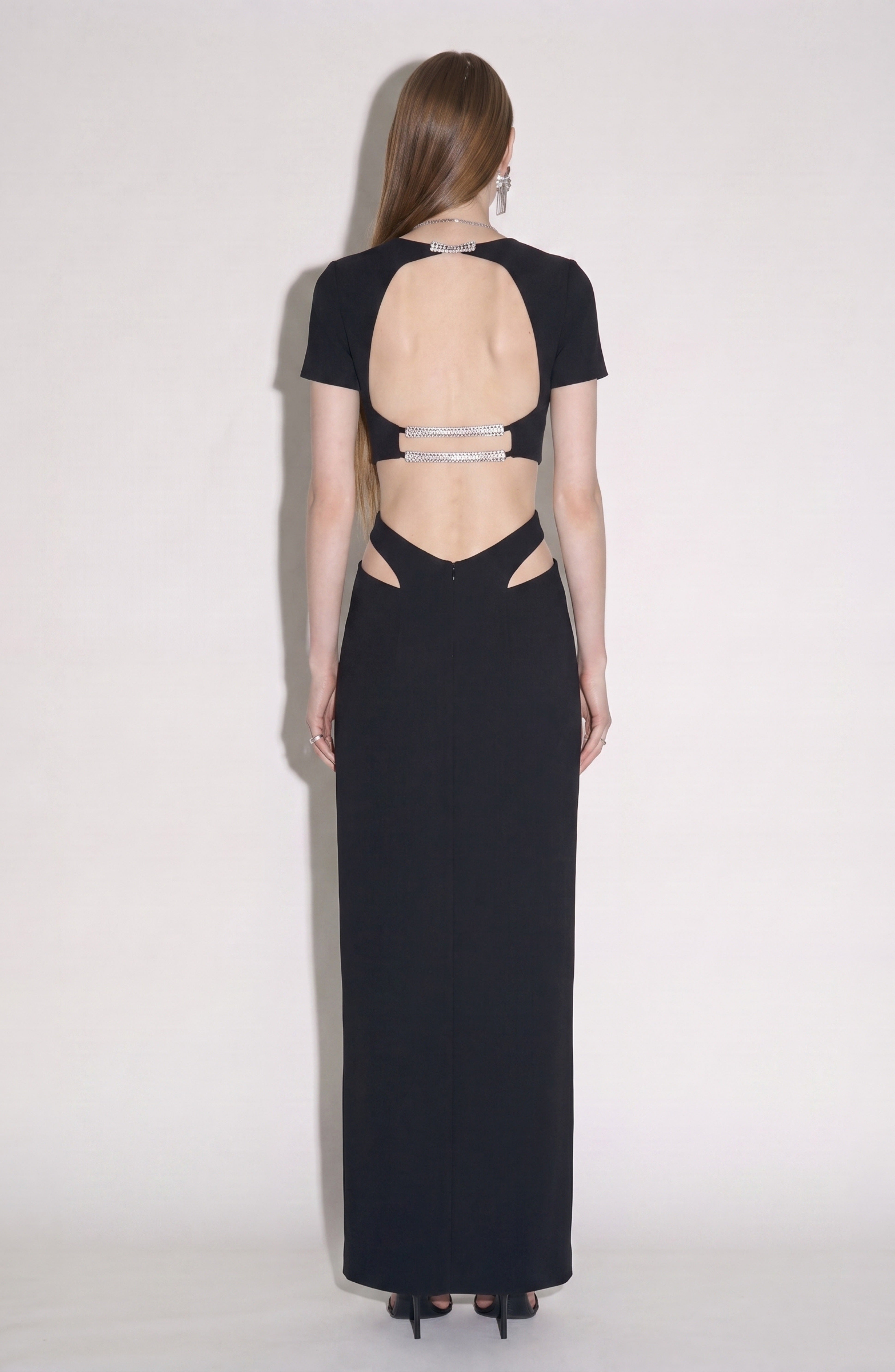 A woman stands with her back to the camera, wearing the LOULOU Crystal-Embellished Cutout Maxi Dress—a sleek black gown featuring waist cutouts and crystal-embellished straps that highlight the open back. Her straight brown hair is worn down, and she completes the look with black high heels.