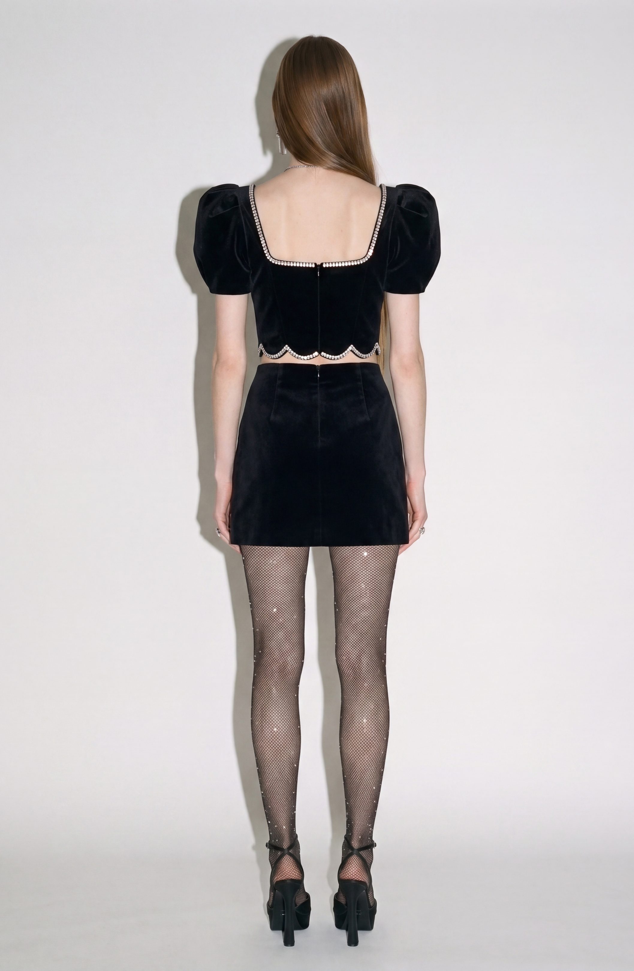 A woman stands facing away, wearing the LOULOU Crystal-Trim Velvet Crop Top paired with a matching black skirt, sparkly sheer tights, and black high-heeled sandals. Her long straight hair cascades down her back as she poses against a plain light background.