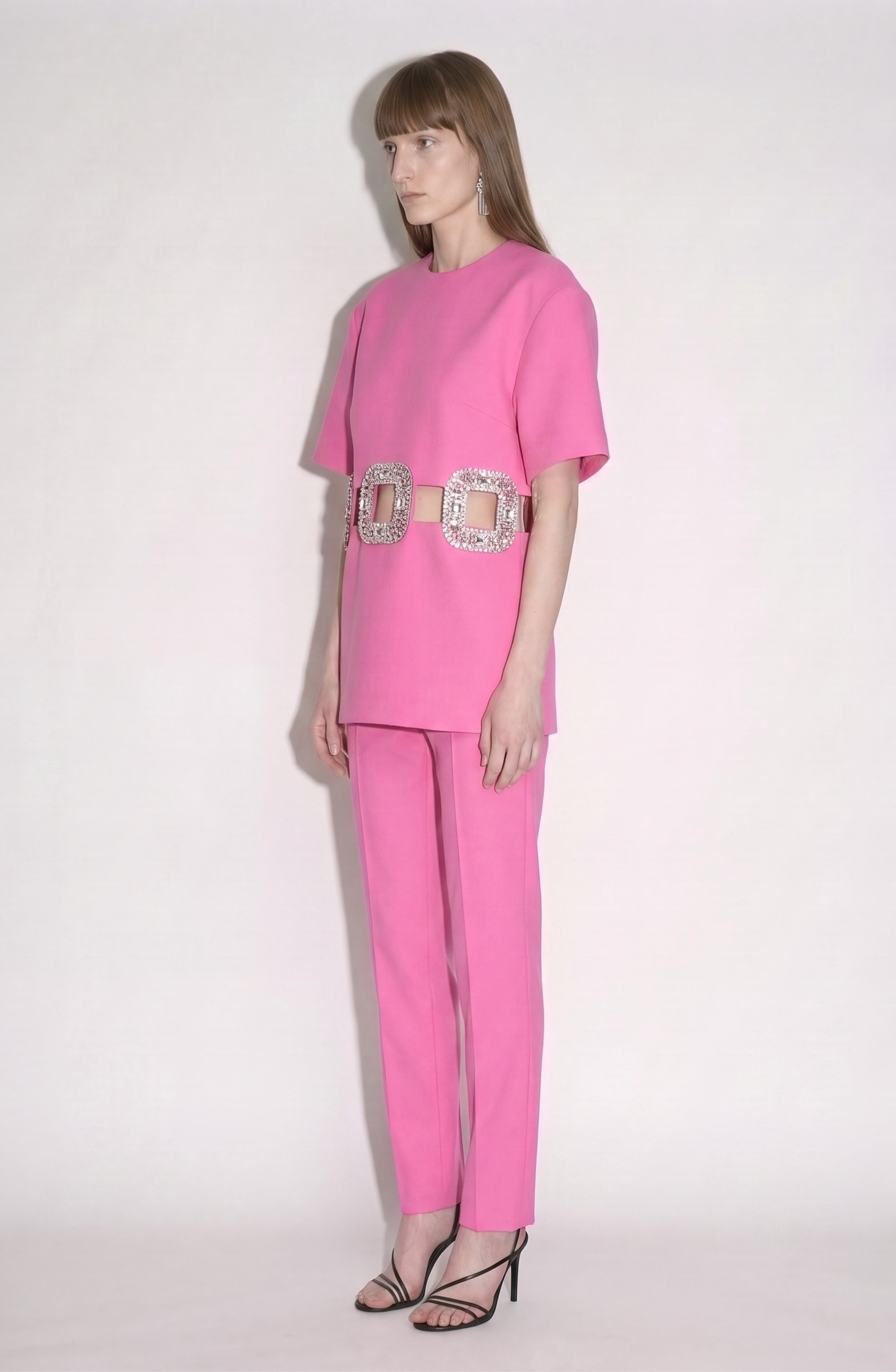 A person stands against a light background wearing the LOULOU Crystal Buckle Cut-Out Top in bright fuchsia, paired with matching pants, a wide belt featuring crystal-embellished buckles, and black heeled sandals.