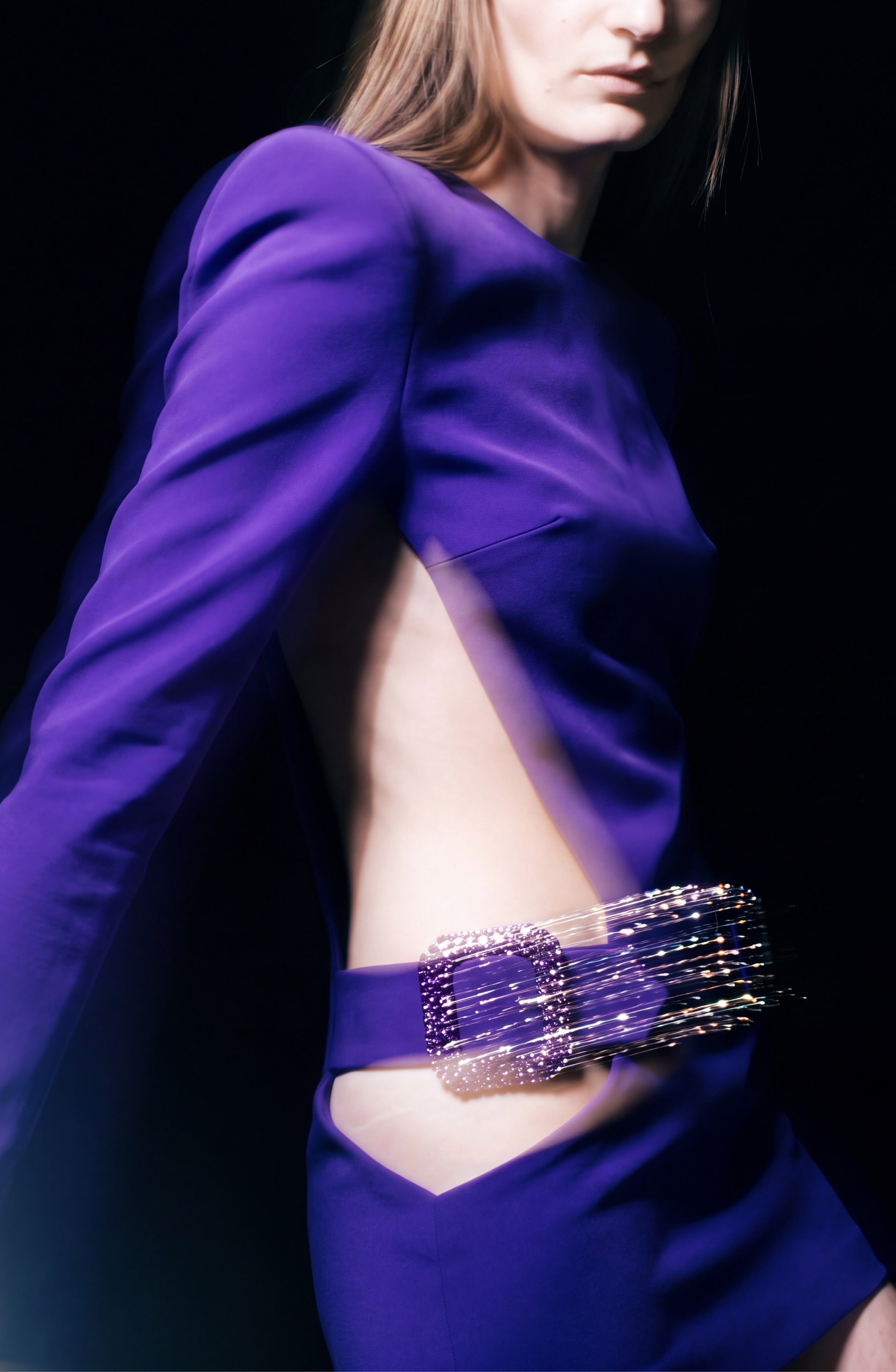A person wearing the LOULOU Crystal Buckle Cutout Mini Dress—a bold purple crepe mini dress featuring structured cut-out sides and a large, sparkling crystal buckle belt—is shown from the side against a dark background.