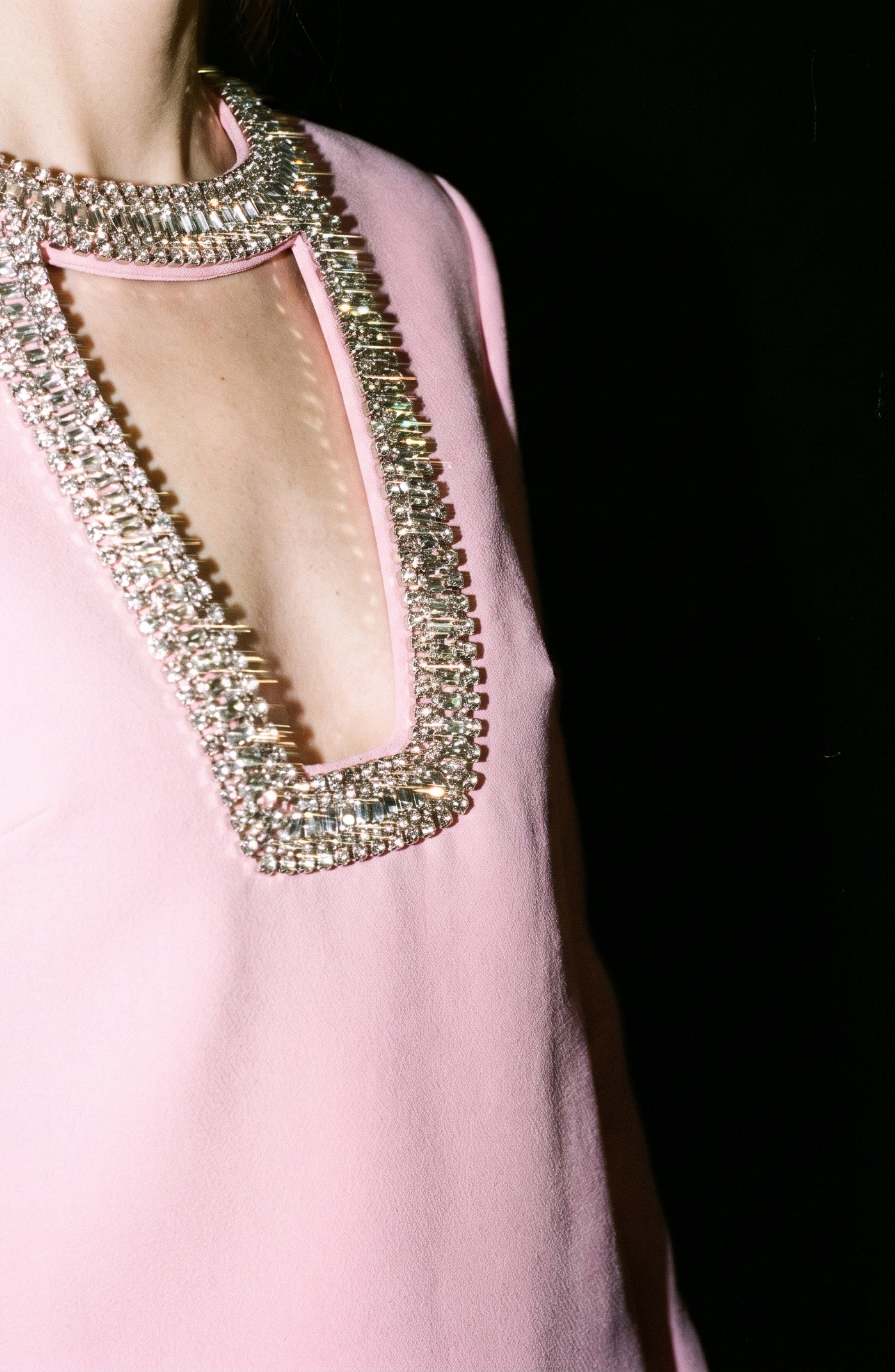 A person wearing the LOULOU Crystal Cutout Mini Dress in pastel pink, featuring a deep square neckline embellished with sparkling rhinestones, stands against a black background.