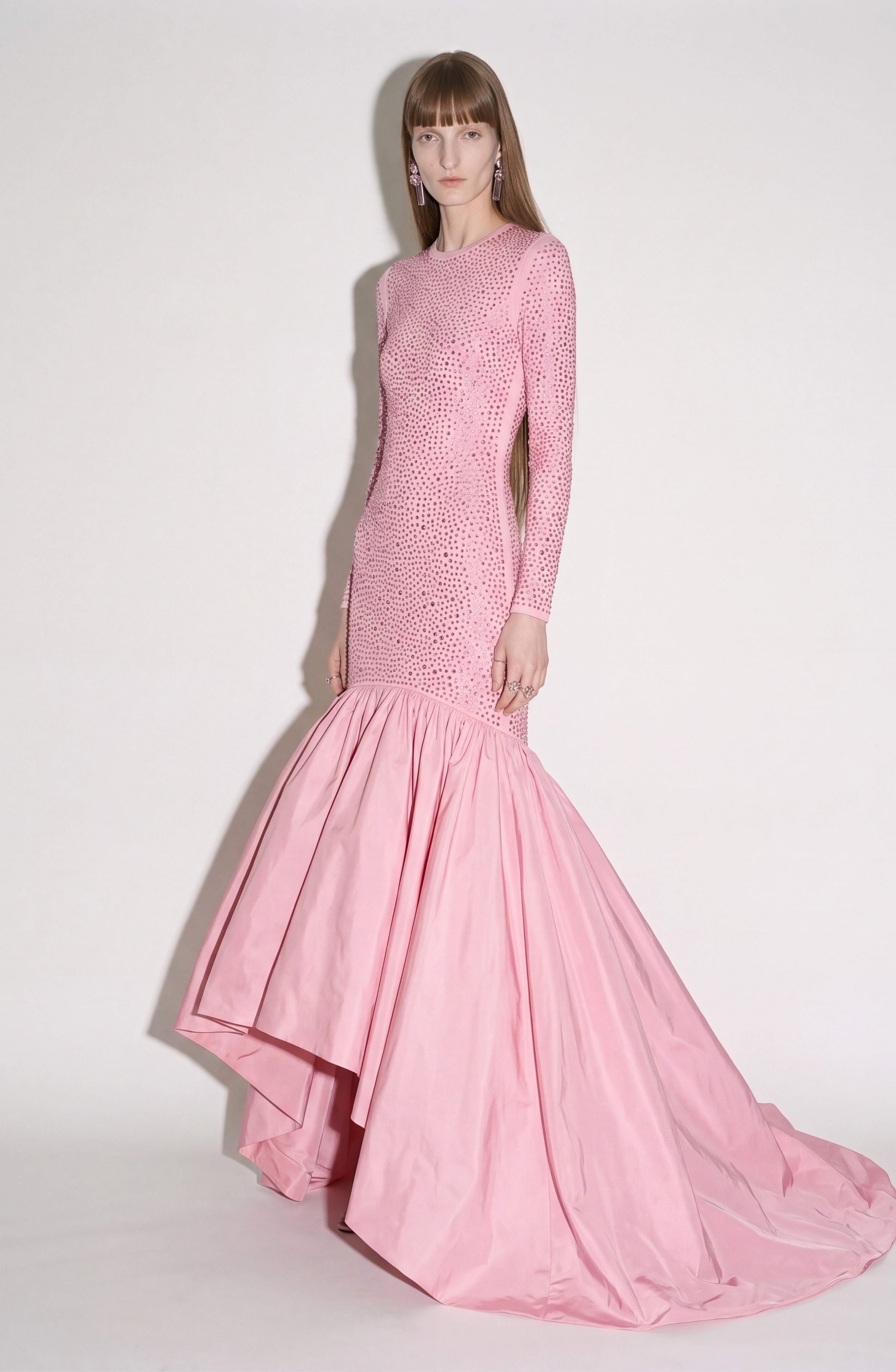 A woman poses against a plain white background, wearing the LOULOU Crystal Embellished High-Low Gown. The dress features long sleeves, a fitted rose pink silhouette, a sparkling crystal-embellished bodice, and a dramatic flared high-low skirt.