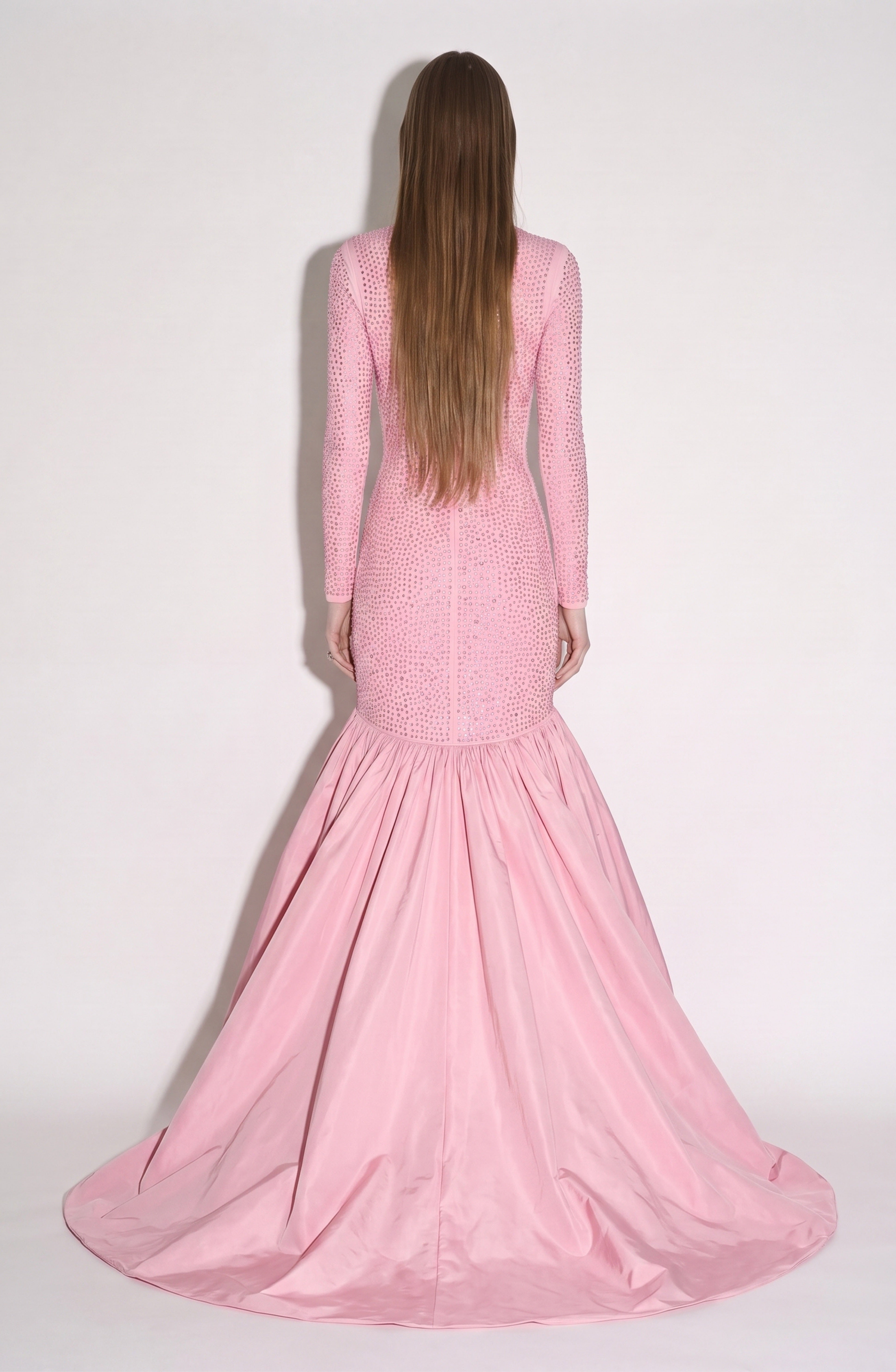 A person with long straight hair, facing away from the camera against a plain light background, wears the LOULOU Crystal Embellished High-Low Gown—a fitted, long-sleeved rose pink dress with a flared taffeta skirt and elegant high-low hem.