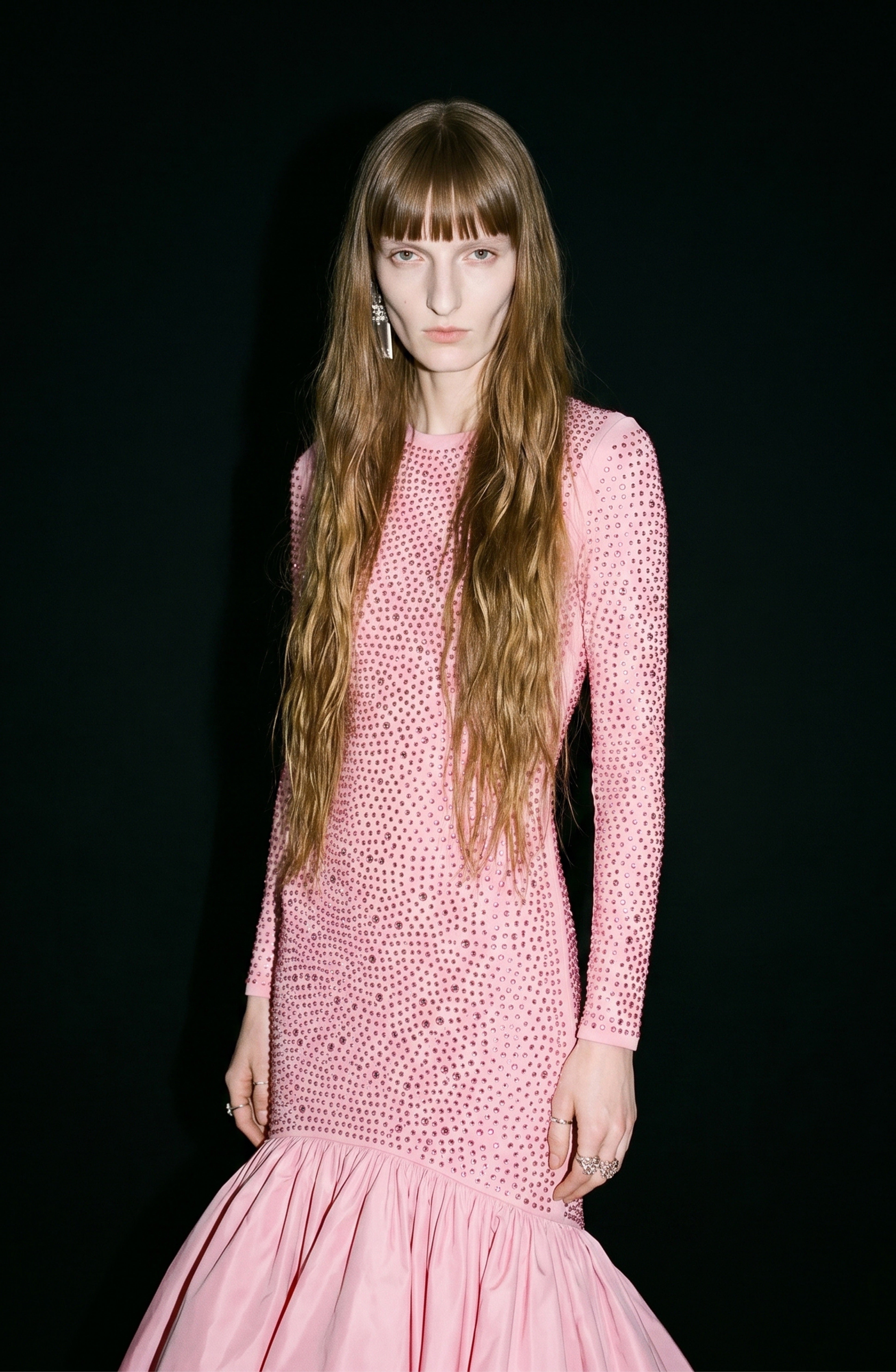 A person with long wavy hair wears the LOULOU Crystal Embellished High-Low Gown, a fitted rose pink dress with a crystal-adorned bodice and a flared hem, standing against a black background.