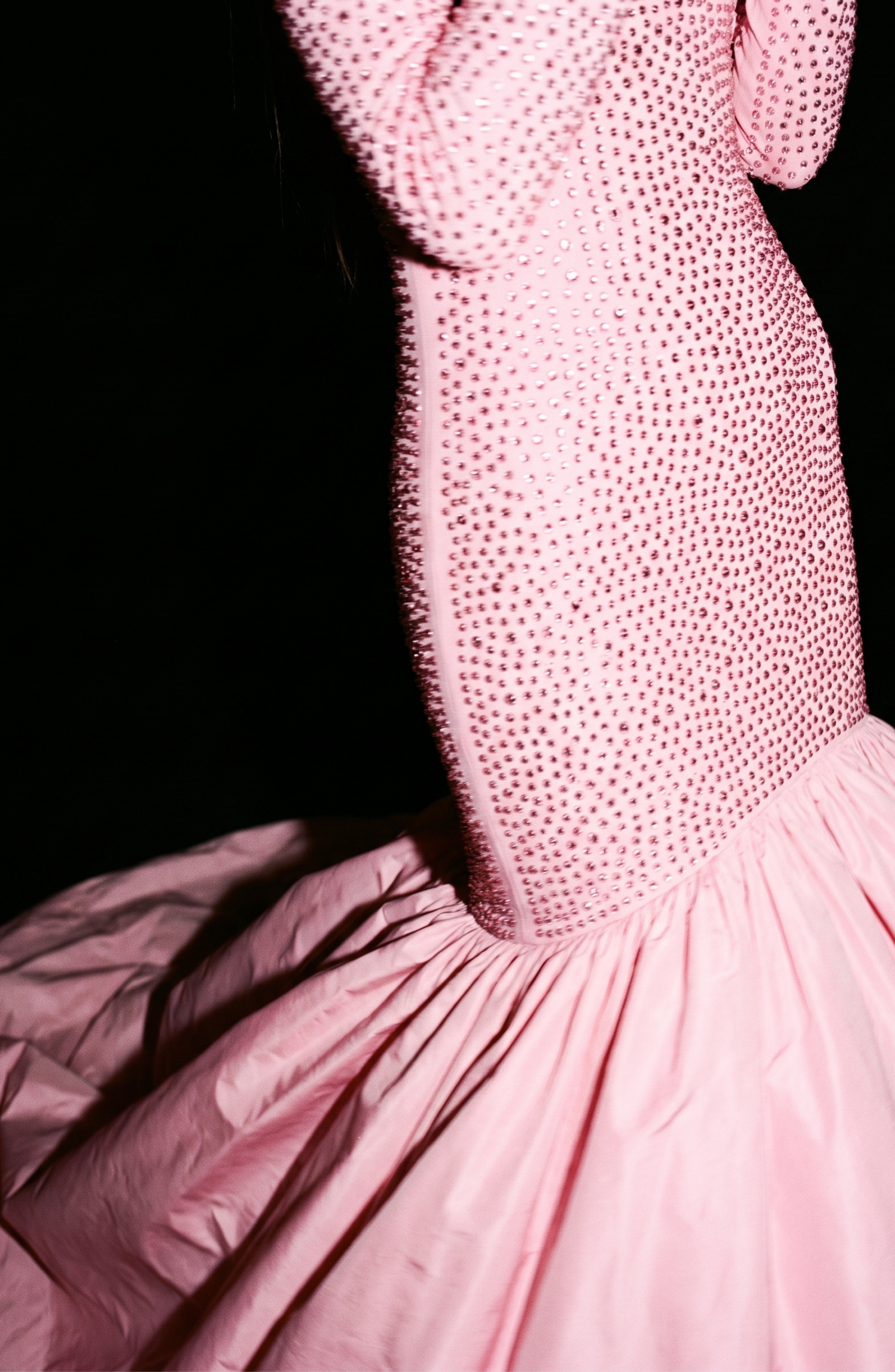 A close-up of LOULOU’s Crystal Embellished High-Low Gown in rose pink, featuring a crystal-embellished bodice and sleeves, set against a black background.