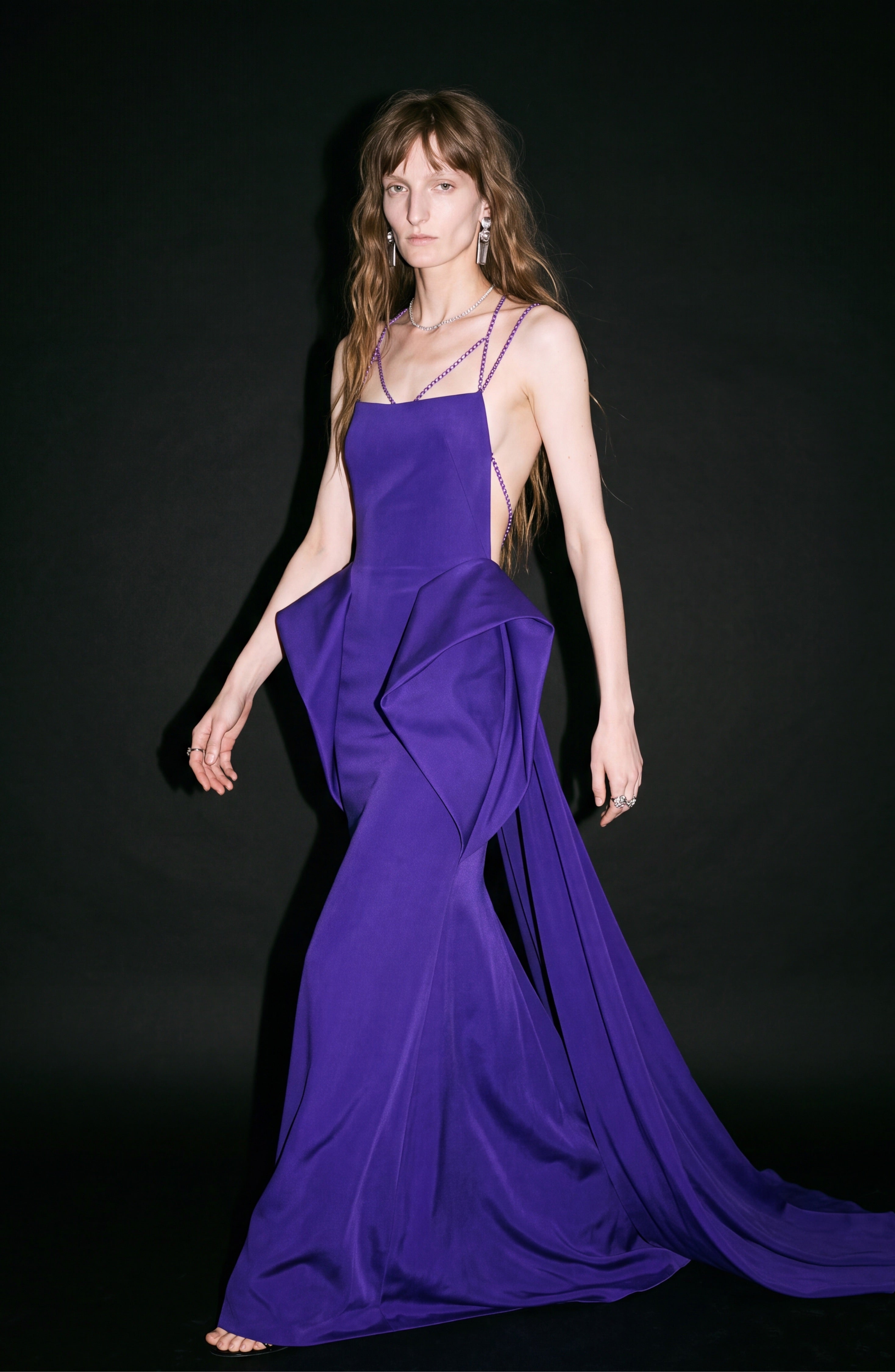 A woman stands against a black background wearing the LOULOU Crystal Strap Peplum Gown, a vibrant purple evening dress featuring geometric side panels, structural peplums, and delicate crystal chain straps.