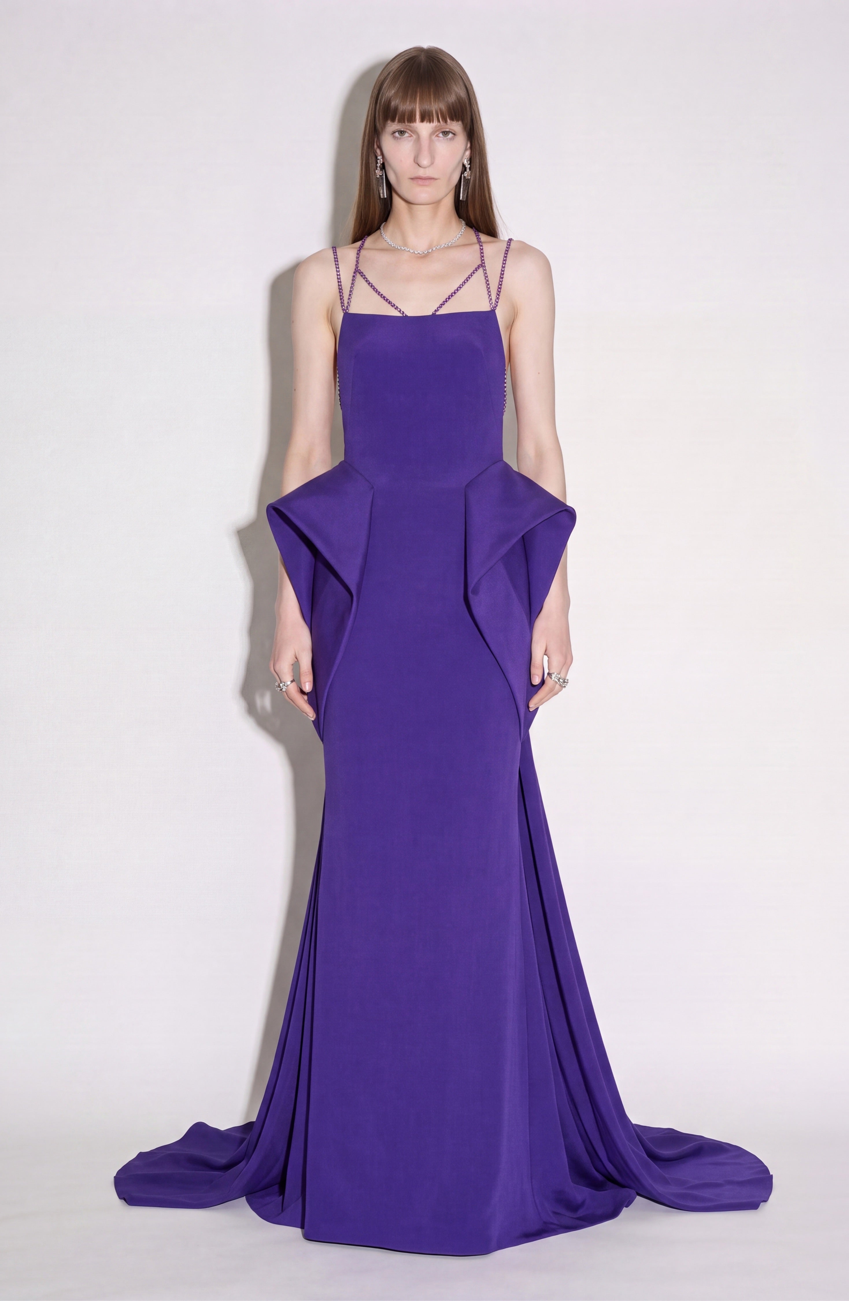 A person wearing the LOULOU Crystal Strap Peplum Gown—a floor-length, sleeveless purple evening gown featuring geometric side panels and subtle structural peplums—stands against a plain light background, facing forward.