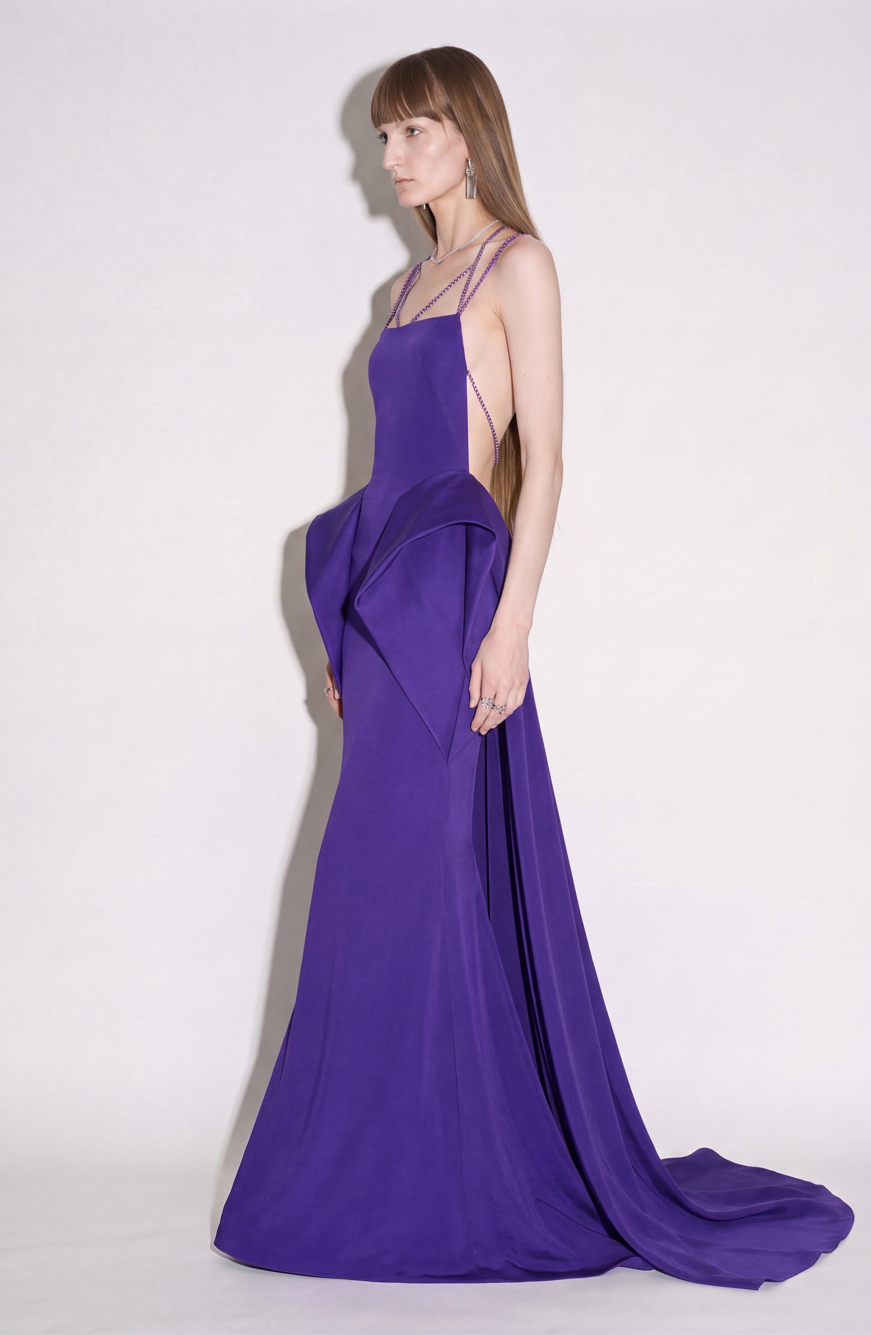 A woman stands against a plain light background, facing left, wearing the LOULOU Crystal Strap Peplum Gown—a sleeveless, backless, floor-length purple evening dress featuring geometric side details and sparkling crystal chain straps.