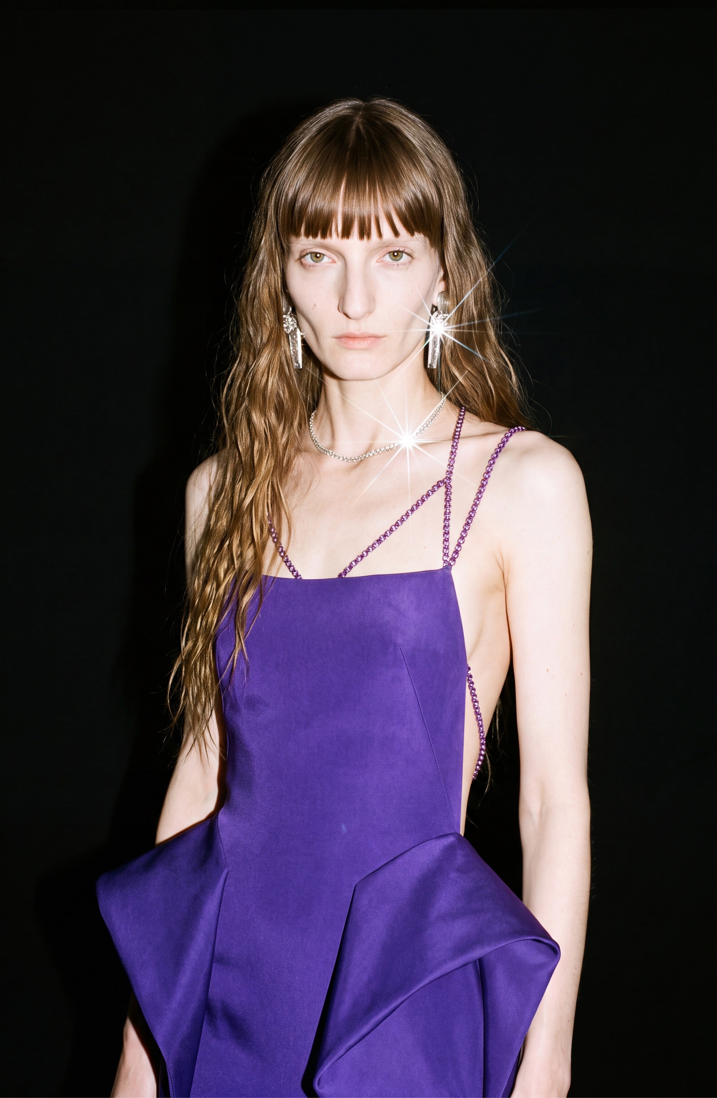 A person with long, wavy brown hair wears the LOULOU Crystal Strap Peplum Gown—a sleeveless purple evening dress with side cutouts—paired with sparkly earrings, standing against a black background.