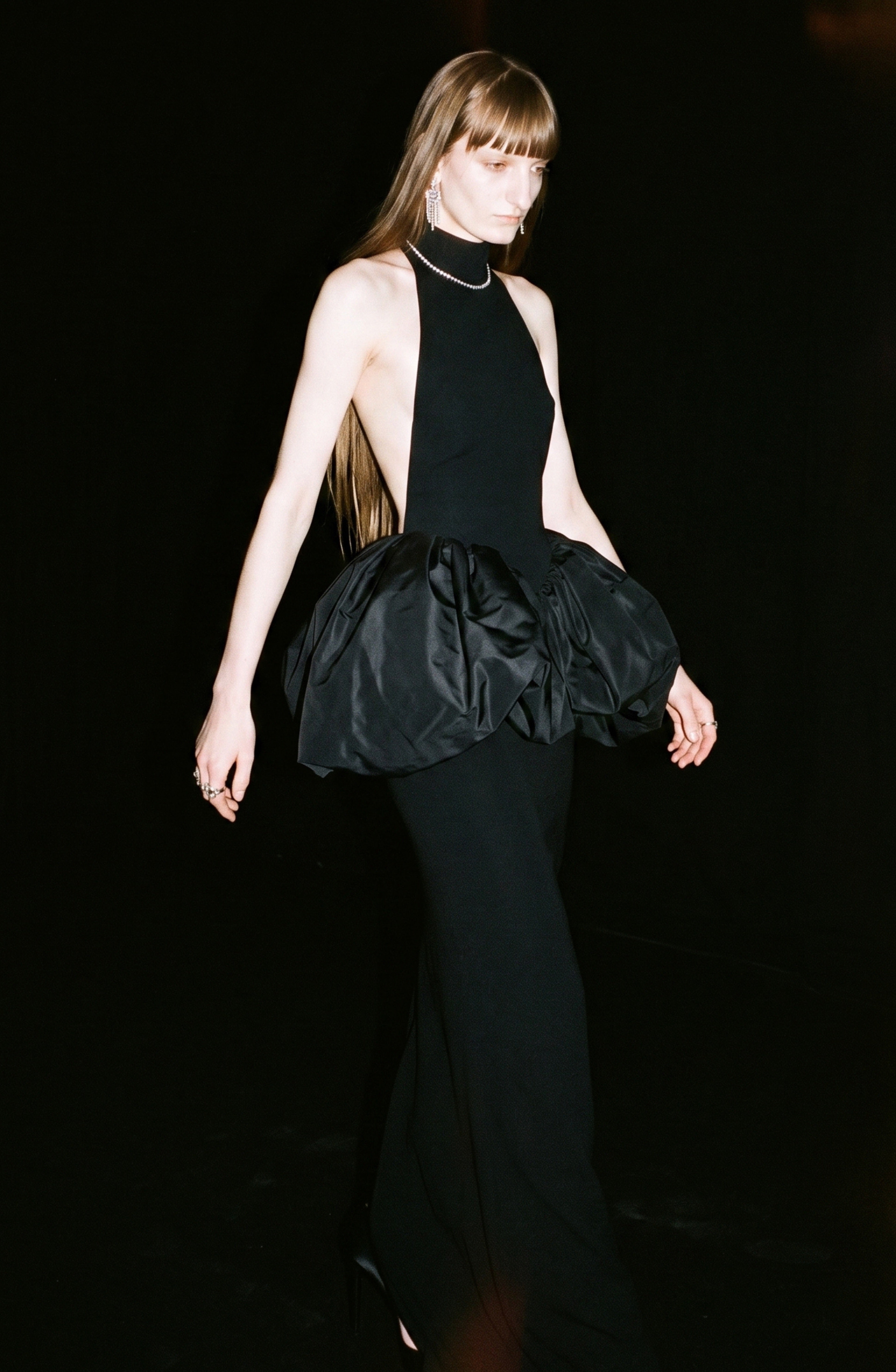 A person with long, straight hair wears the LOULOU Crystal Trim Peplum Maxi Dress—a sleeveless black maxi dress featuring a high neck and dramatic, voluminous ruffles at the waist—while standing against a dark background.