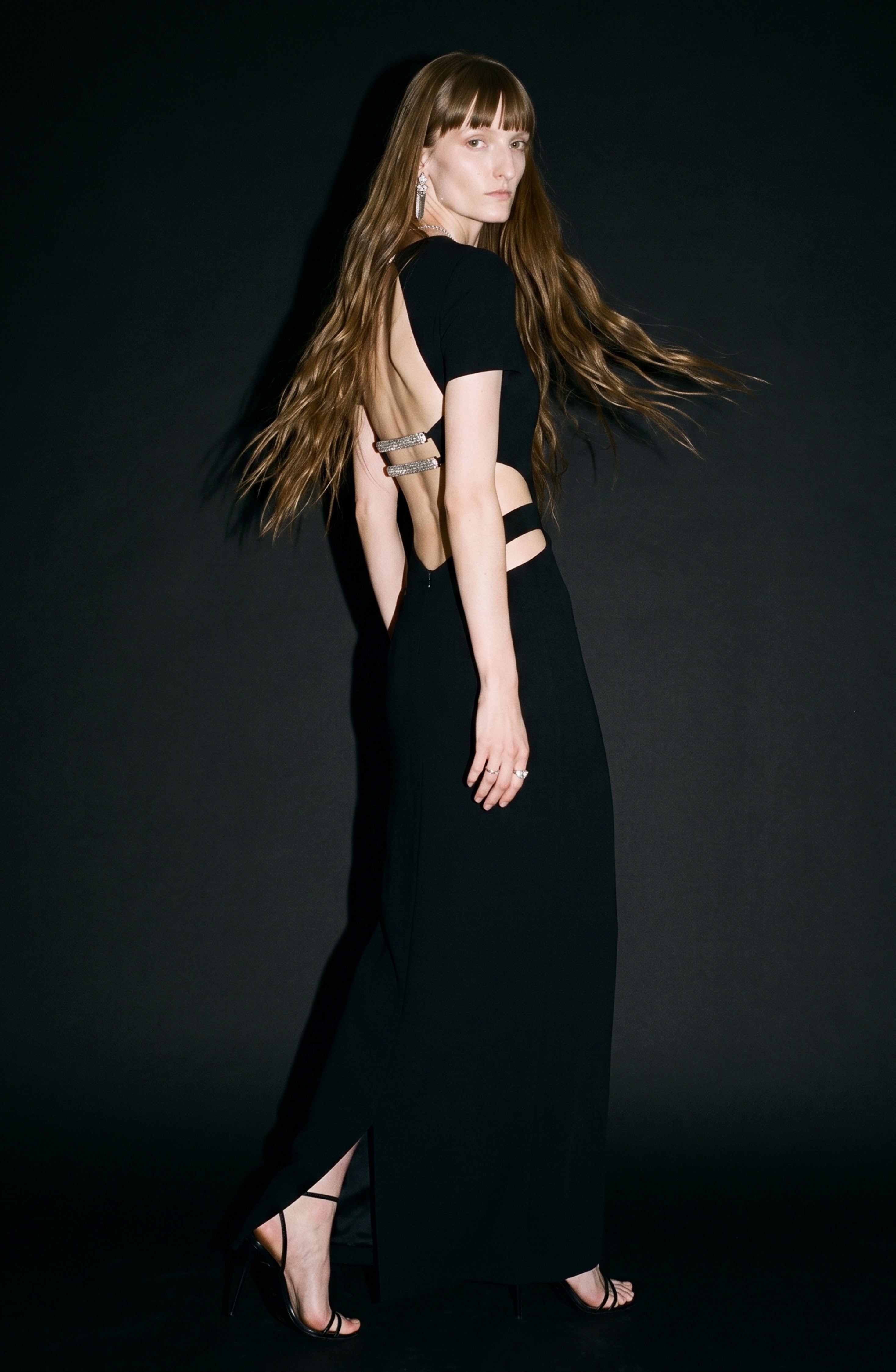 A woman with long brown hair wears the LOULOU Cut-Out Maxi Dress in jet black, distinguished by waist cut-outs and crystal-embellished straps, paired with black heels. She stands against a dark background, turning slightly with one foot forward as her hair flows gracefully.