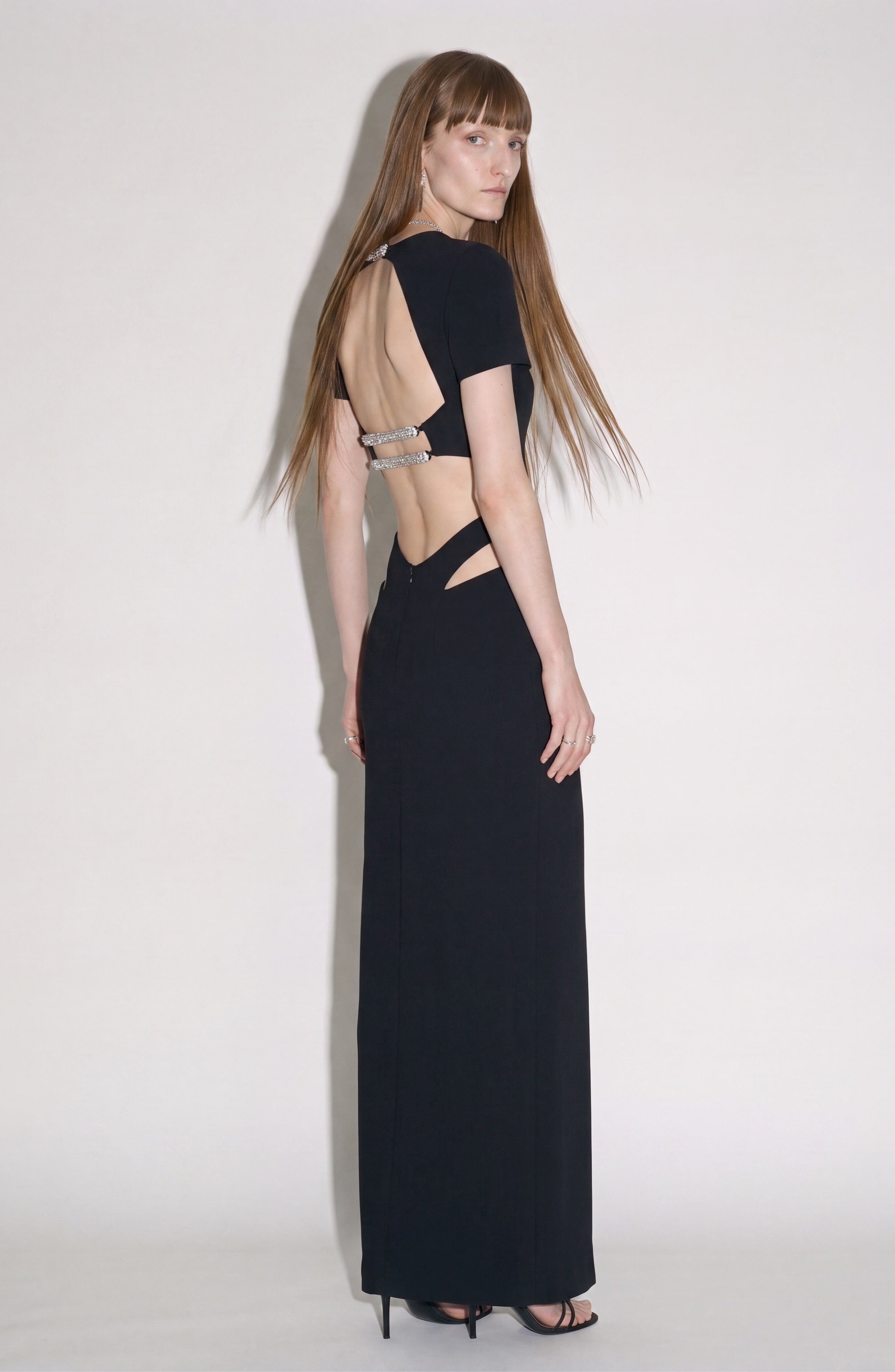 A woman stands sideways, looking over her shoulder, wearing the LOULOU Cut-Out Maxi Dress—an elegant jet black maxi dress featuring waist cut-outs and crystal-embellished straps. She has long straight hair and pairs the look with black high-heeled shoes.