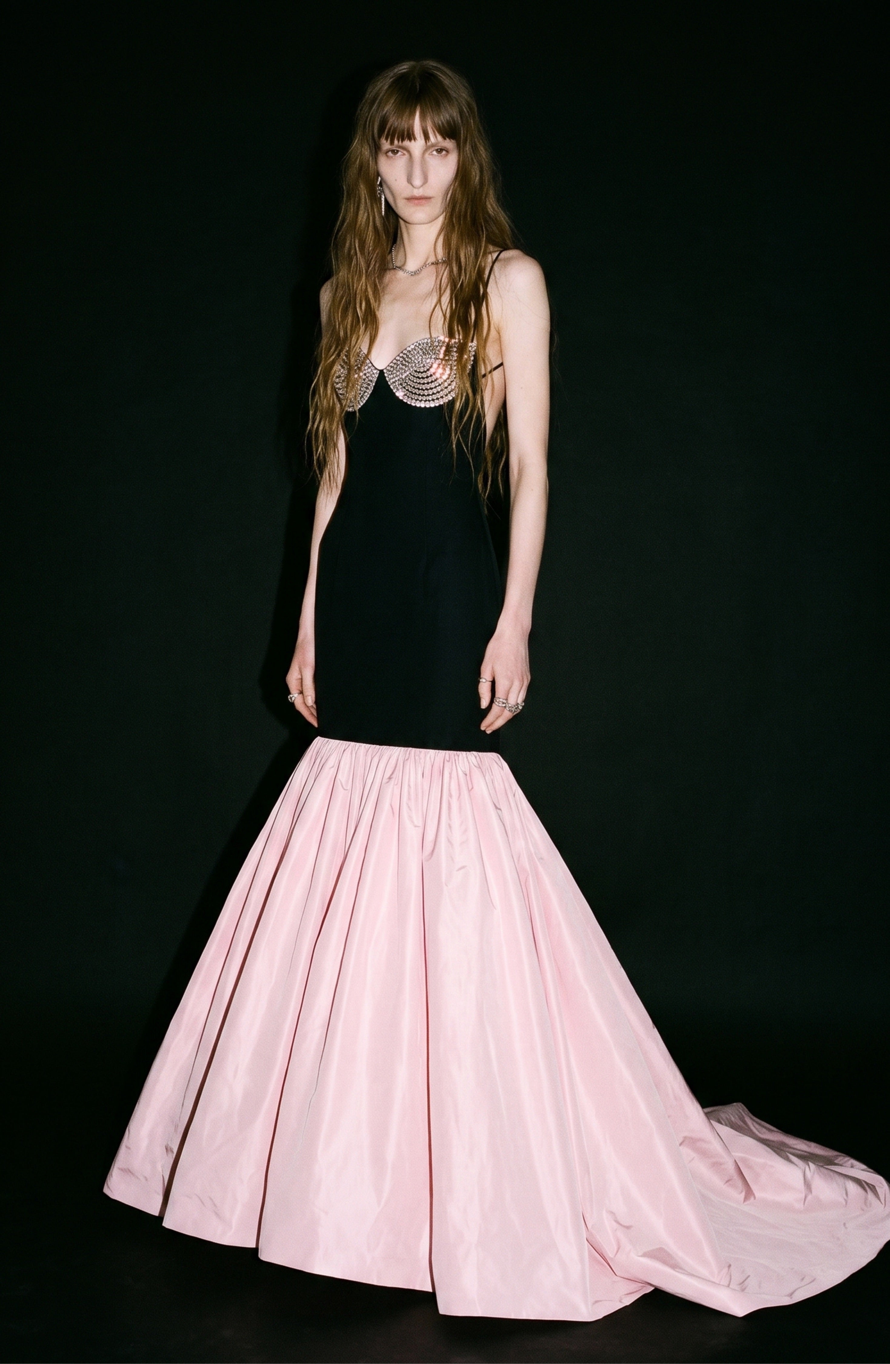 A woman stands against a plain black background wearing the LOULOU Detailed Bustier Mermaid Gown, featuring a fitted black silhouette with a crystal-embellished bodice and a flared pink taffeta skirt.