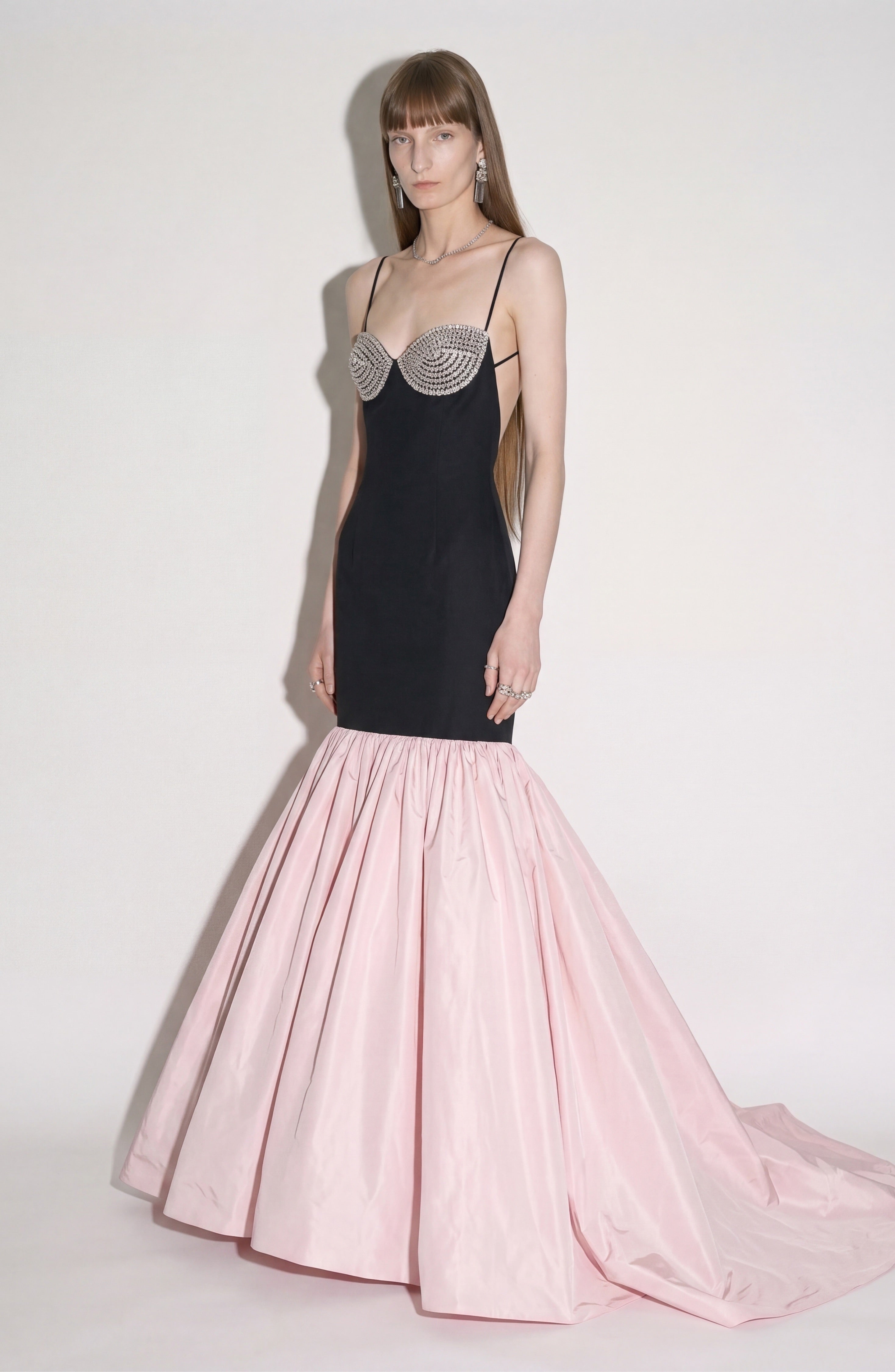 A woman stands against a plain light background wearing the LOULOU Detailed Bustier Mermaid Gown, featuring thin straps, a crystal-embellished bodice, and a voluminous light pink taffeta mermaid skirt.