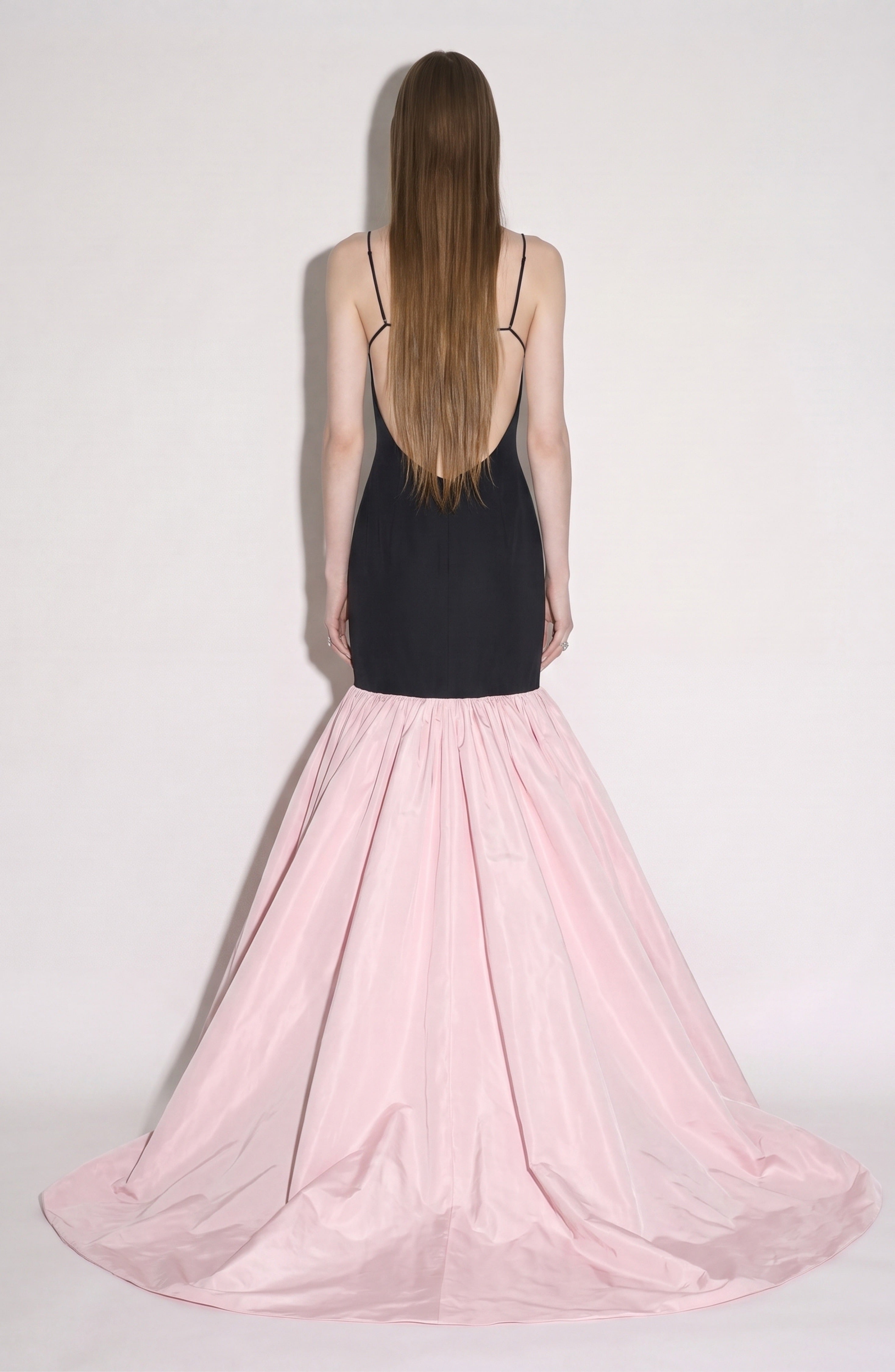 A person with long straight hair faces away, wearing the LOULOU Detailed Bustier Mermaid Gown: a black backless dress with thin straps and a dramatic, floor-length taffeta skirt that flares out at the bottom.