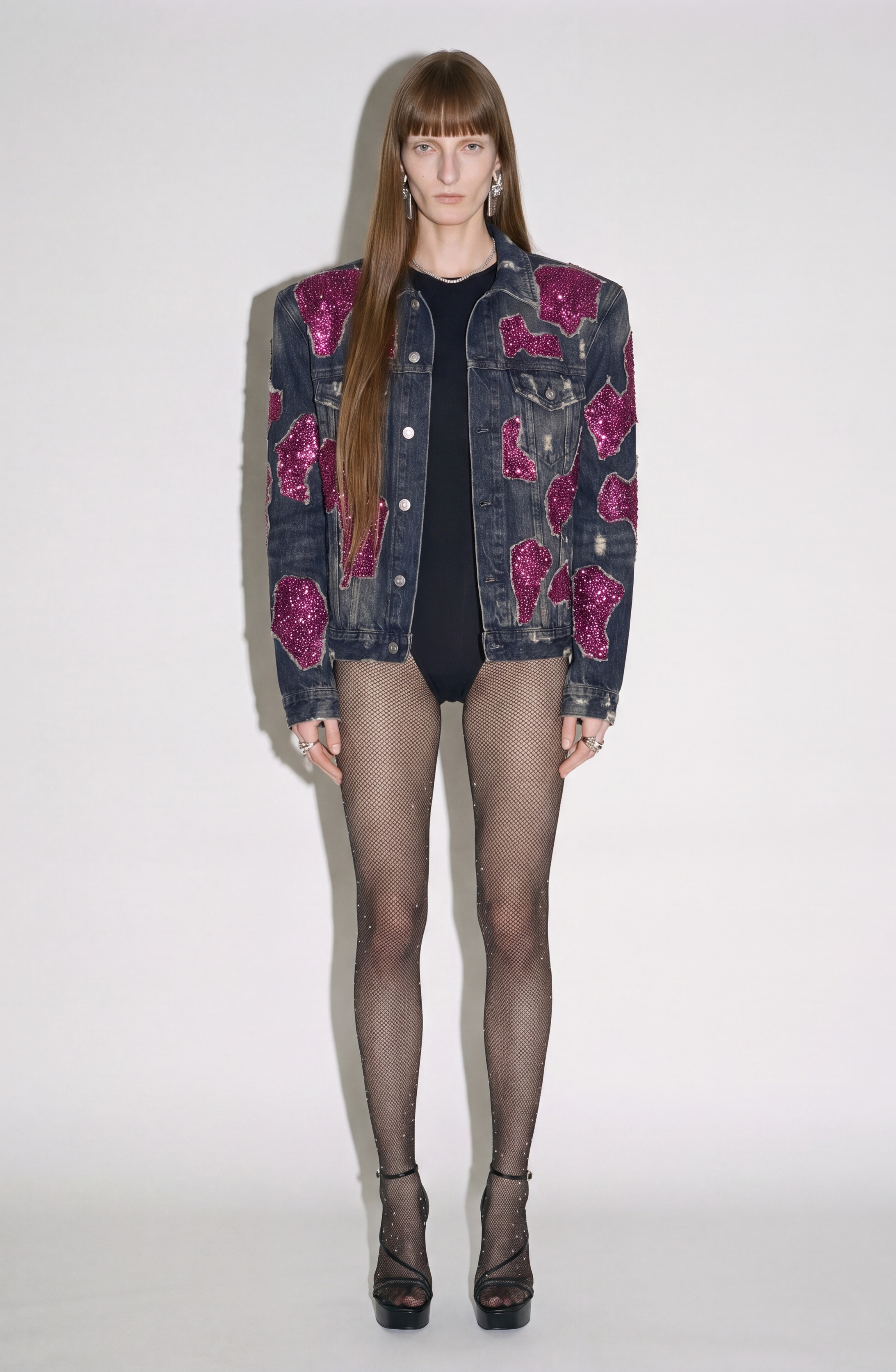 A person stands against a white background, wearing the LOULOU Embellished Denim Jacket with magenta sequin appliqués, paired with a black bodysuit, sheer embellished fishnet tights, and black high-heeled sandals.