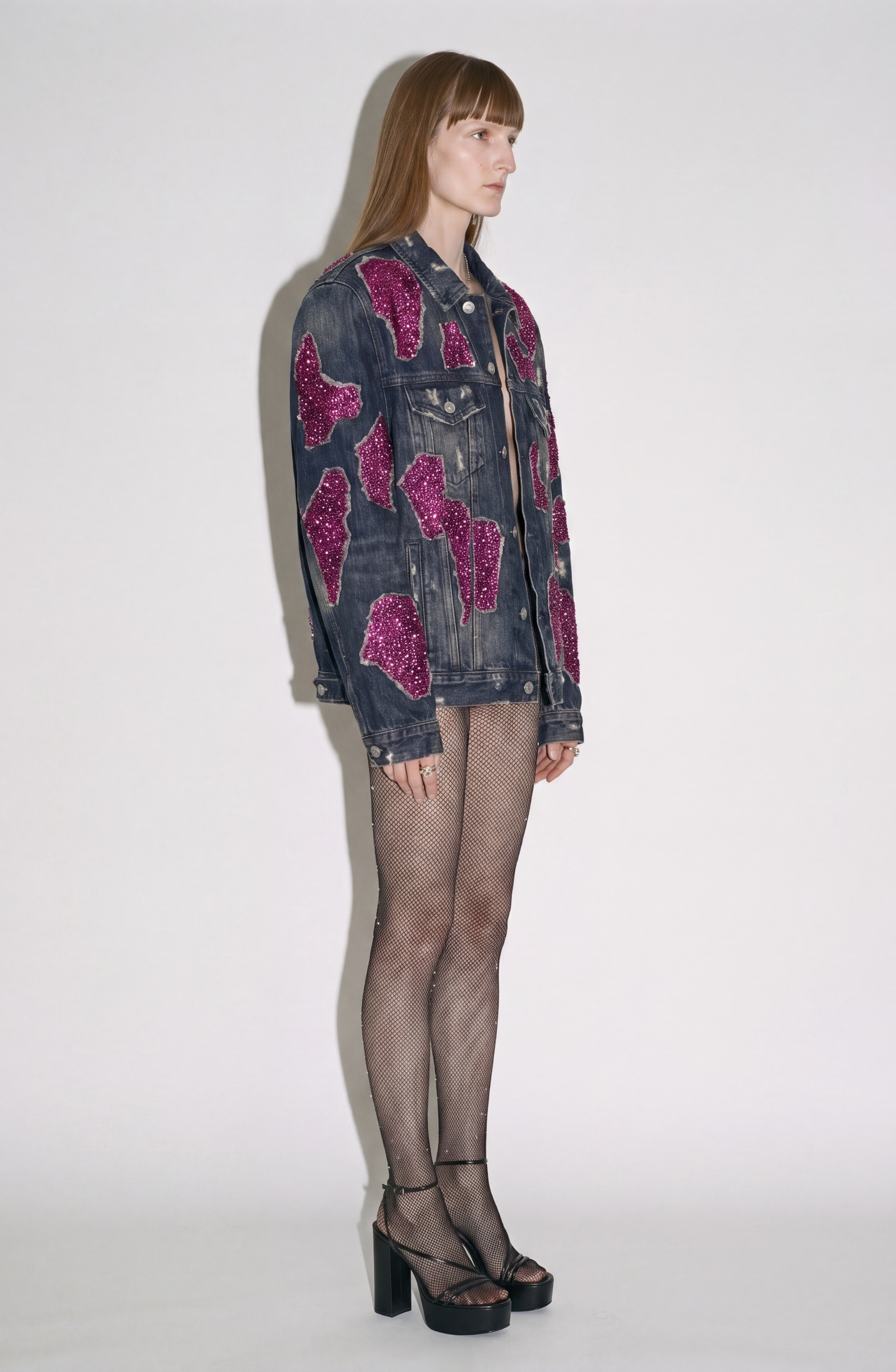 A person stands sideways against a plain white background, wearing the LOULOU Embellished Denim Jacket, featuring vintage-washed denim and magenta sequin appliqués, styled with fishnet tights and black platform heels.