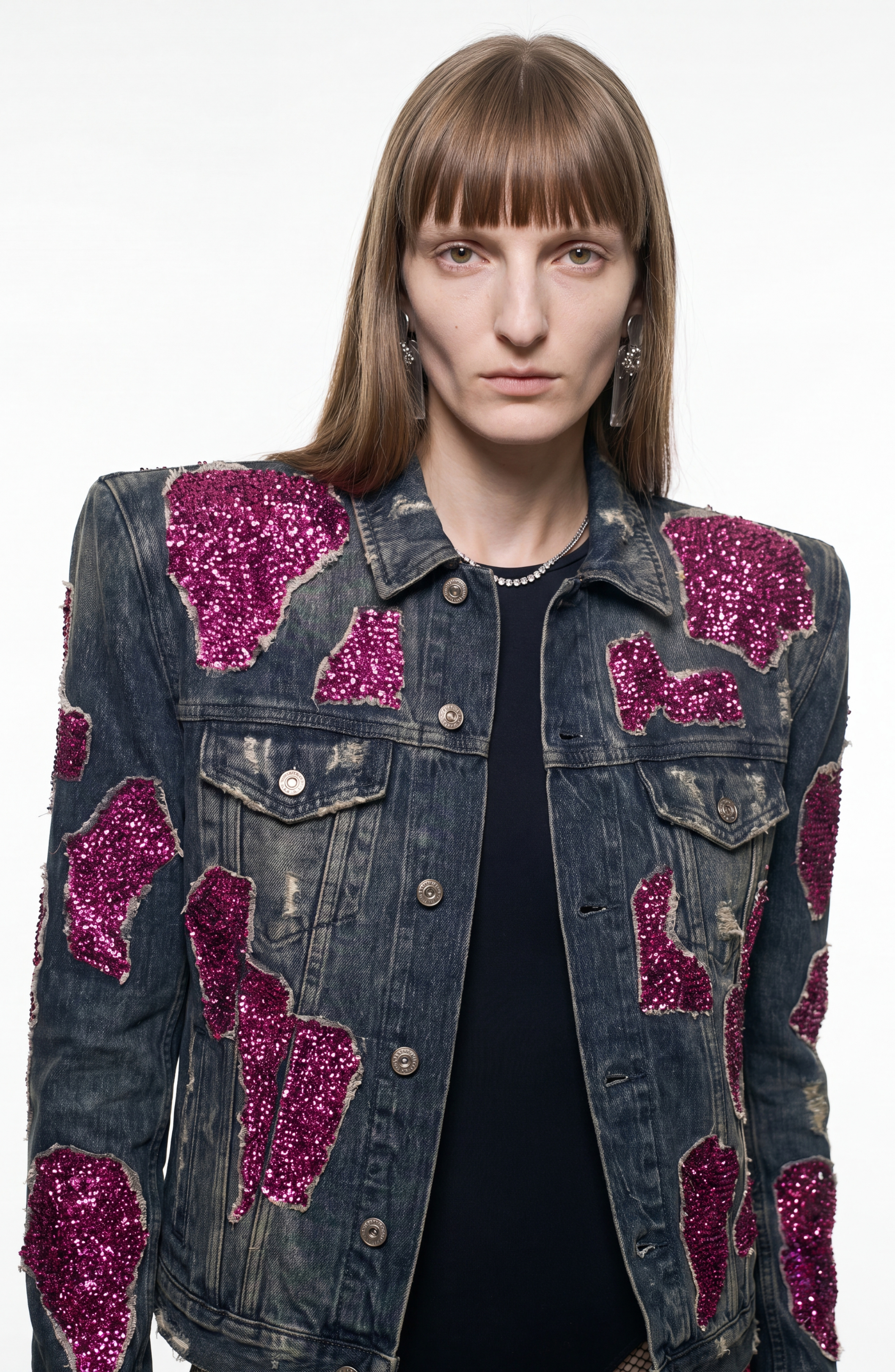 A person with straight brown hair wears the LOULOU Embellished Denim Jacket, featuring a vintage wash and magenta sequin appliqués, styled with a black shirt, necklace, and earrings, standing against a plain white background.