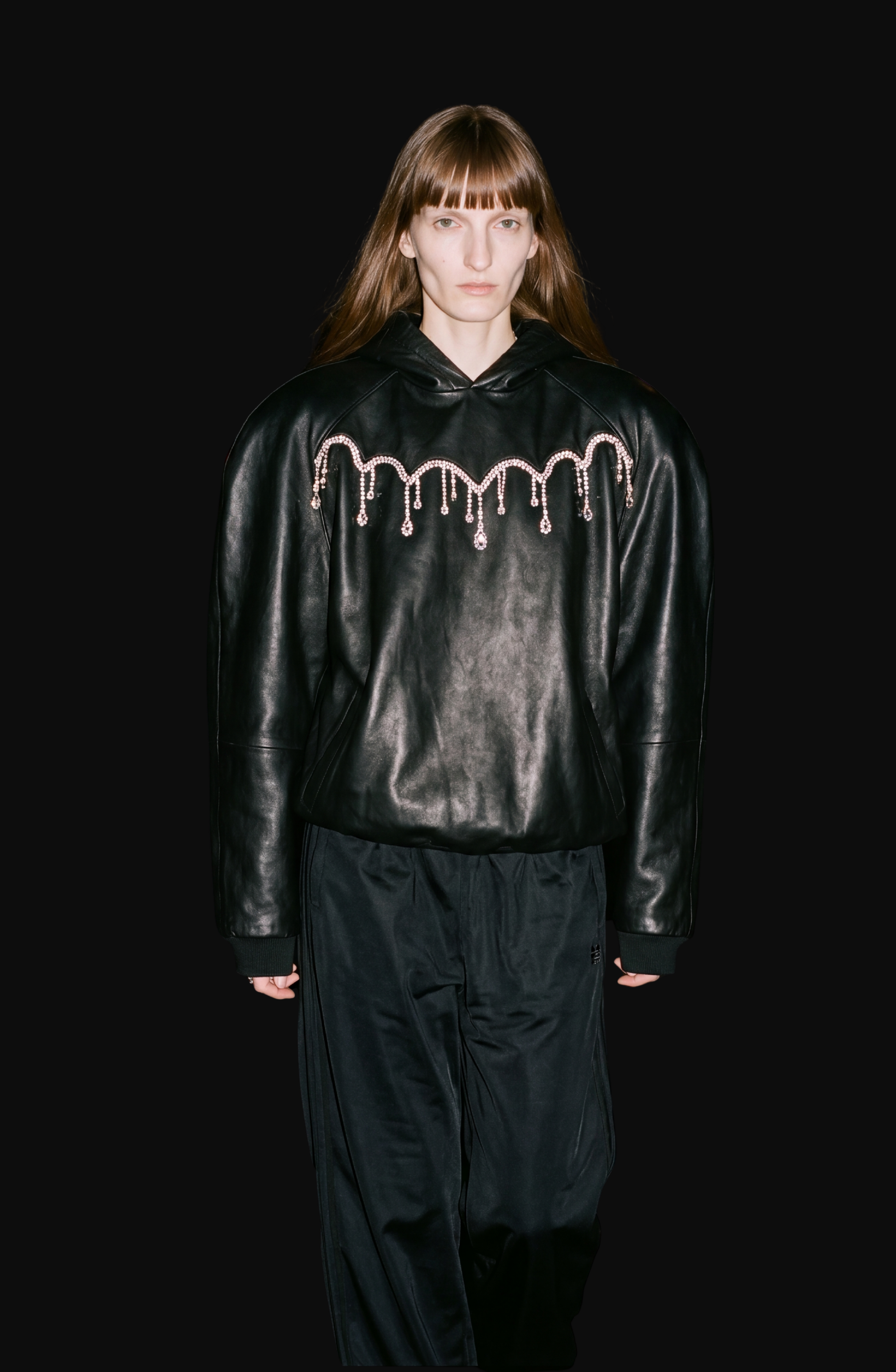 A person with long brown hair wears the LOULOU Embellished Leather Hoodie—an oversized black leather piece adorned with crystal chandelier-inspired pink gem-like chains—paired with wide black pants, standing against a solid black background.