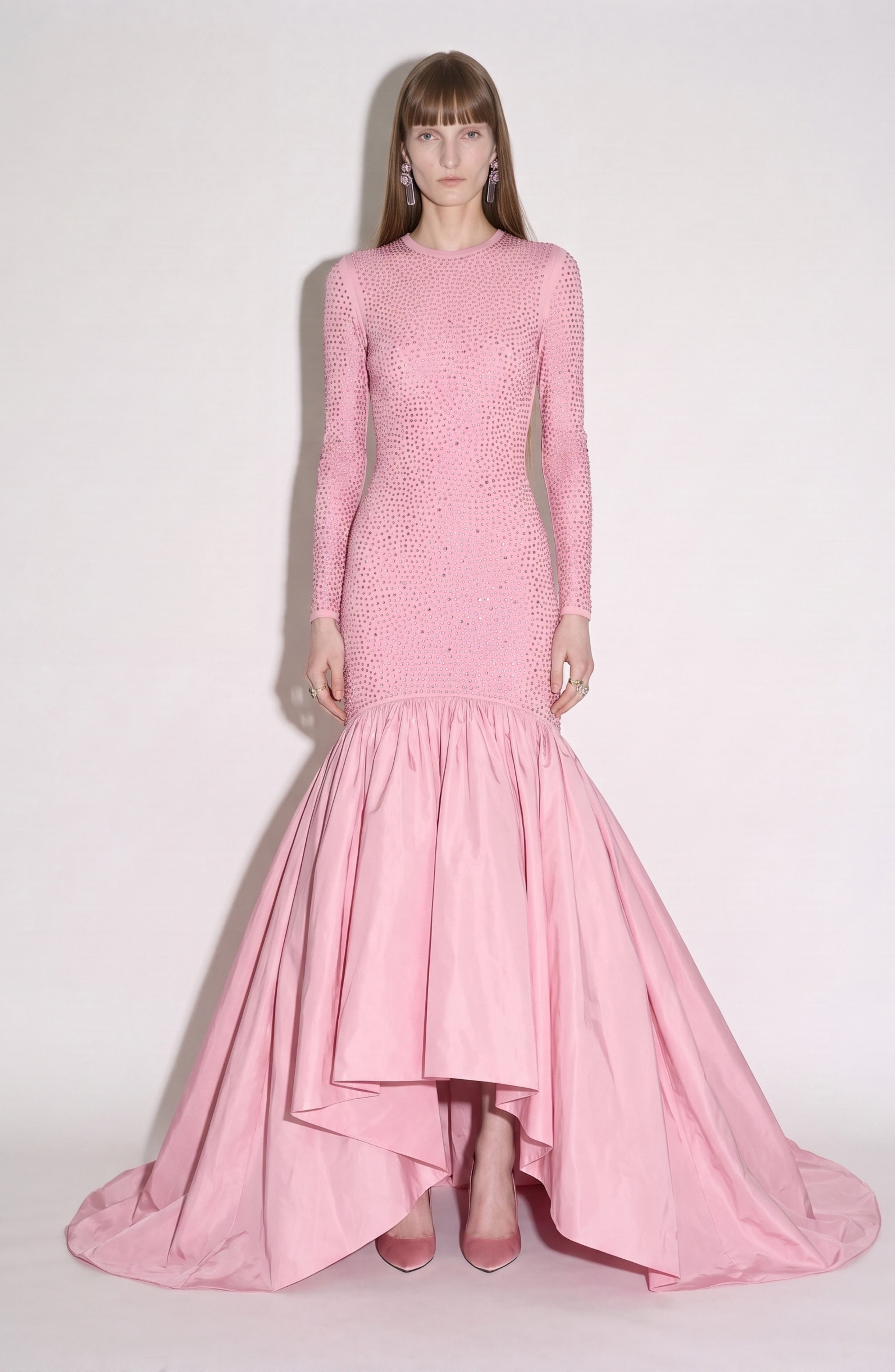 A person stands against a plain background wearing the LOULOU Embellished Taffeta Gown, a long, light pink evening dress featuring fitted long sleeves, sparkling crystal-embellished details, and a flared hem, paired with matching pink heels.