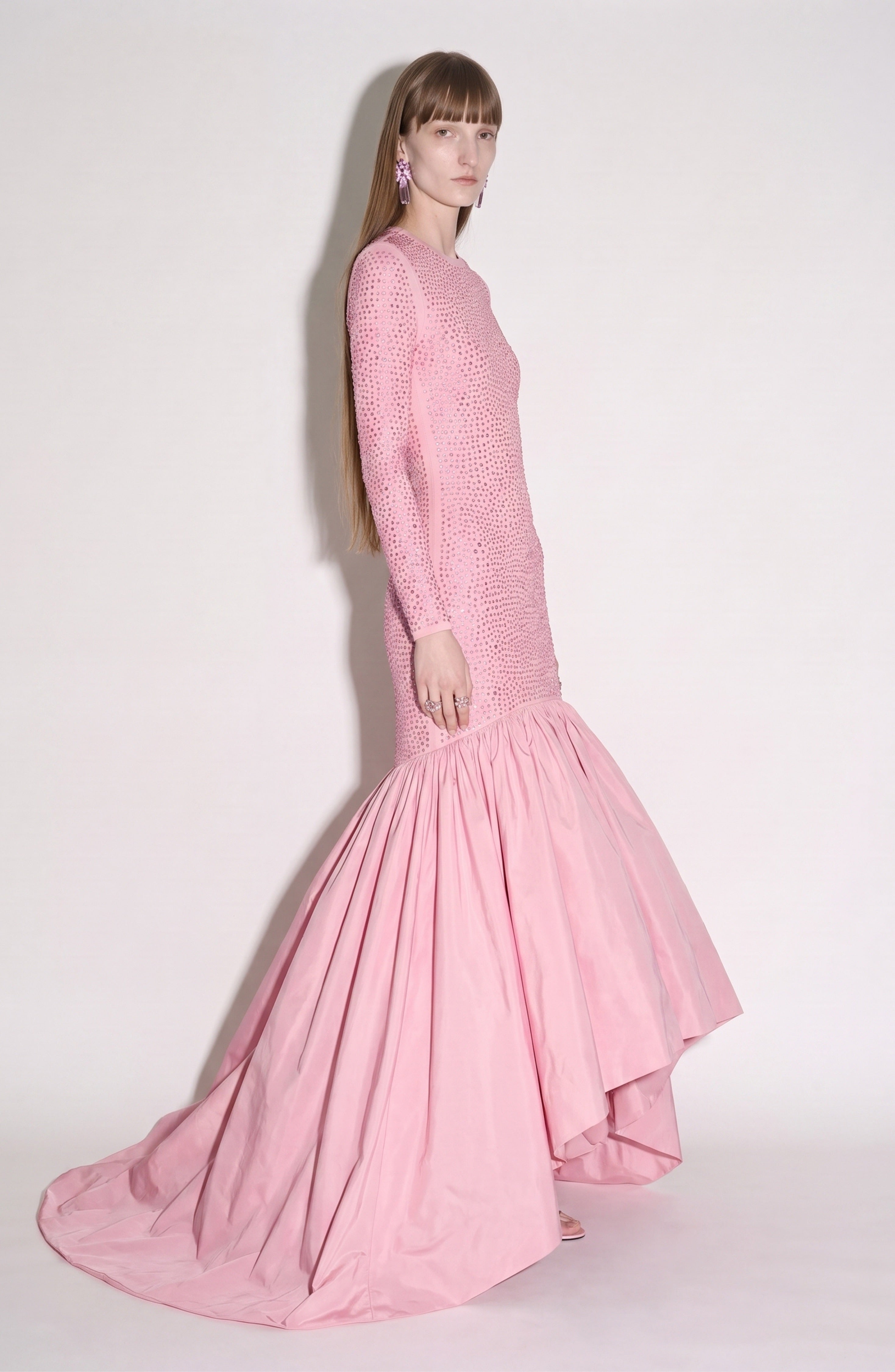 A person with long hair stands sideways against a plain light background, wearing the LOULOU Embellished Taffeta Gown—a fitted, long-sleeved pink evening dress featuring textured detailing and a dramatic mermaid-style skirt.