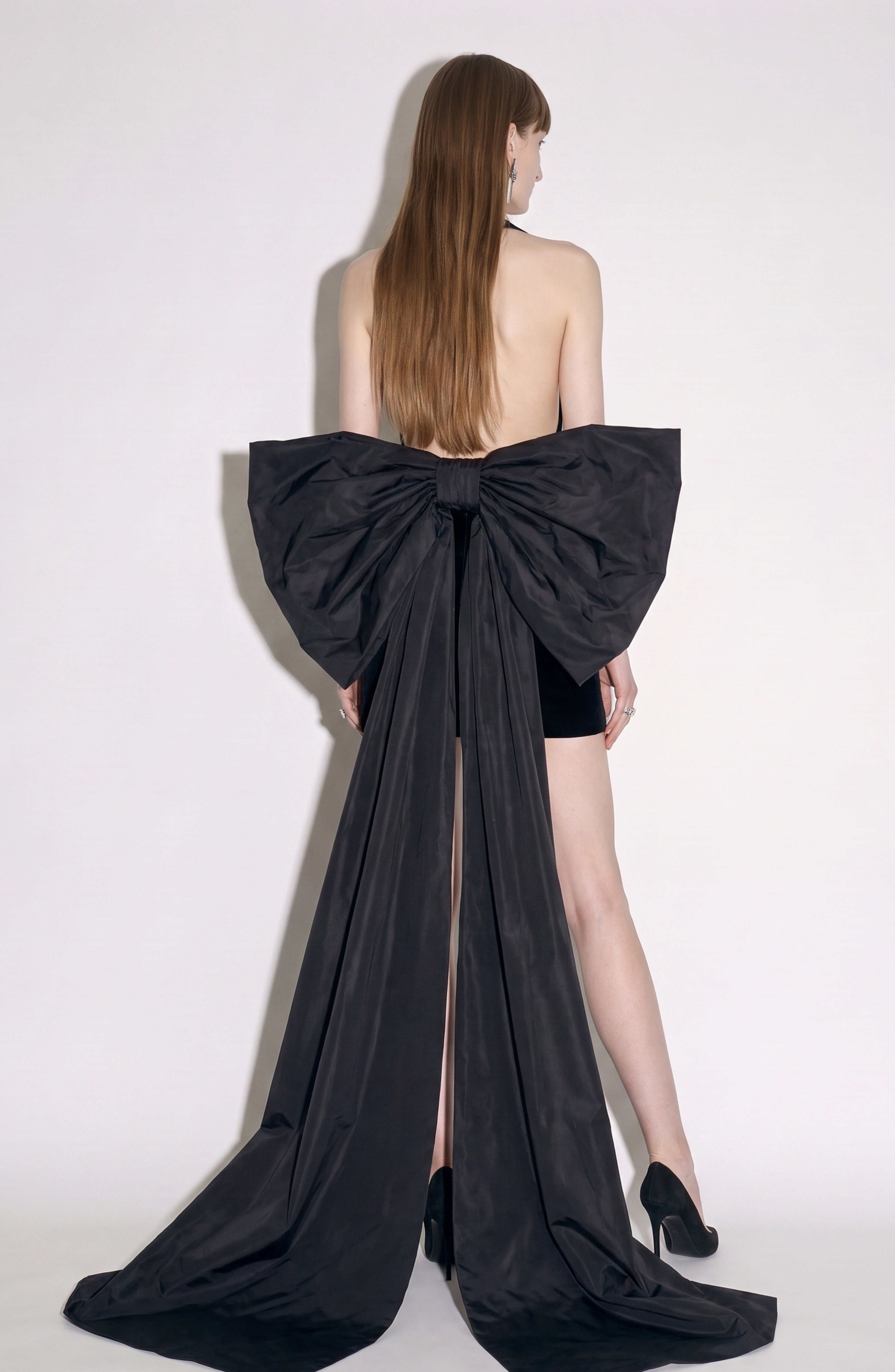 A woman stands facing away, wearing the LOULOU Velvet Bow Mini Dress—a striking black velvet mini dress featuring a dramatic oversized taffeta bow and long train—paired with black heels. Her straight, long hair cascades down her back against a simple light background.