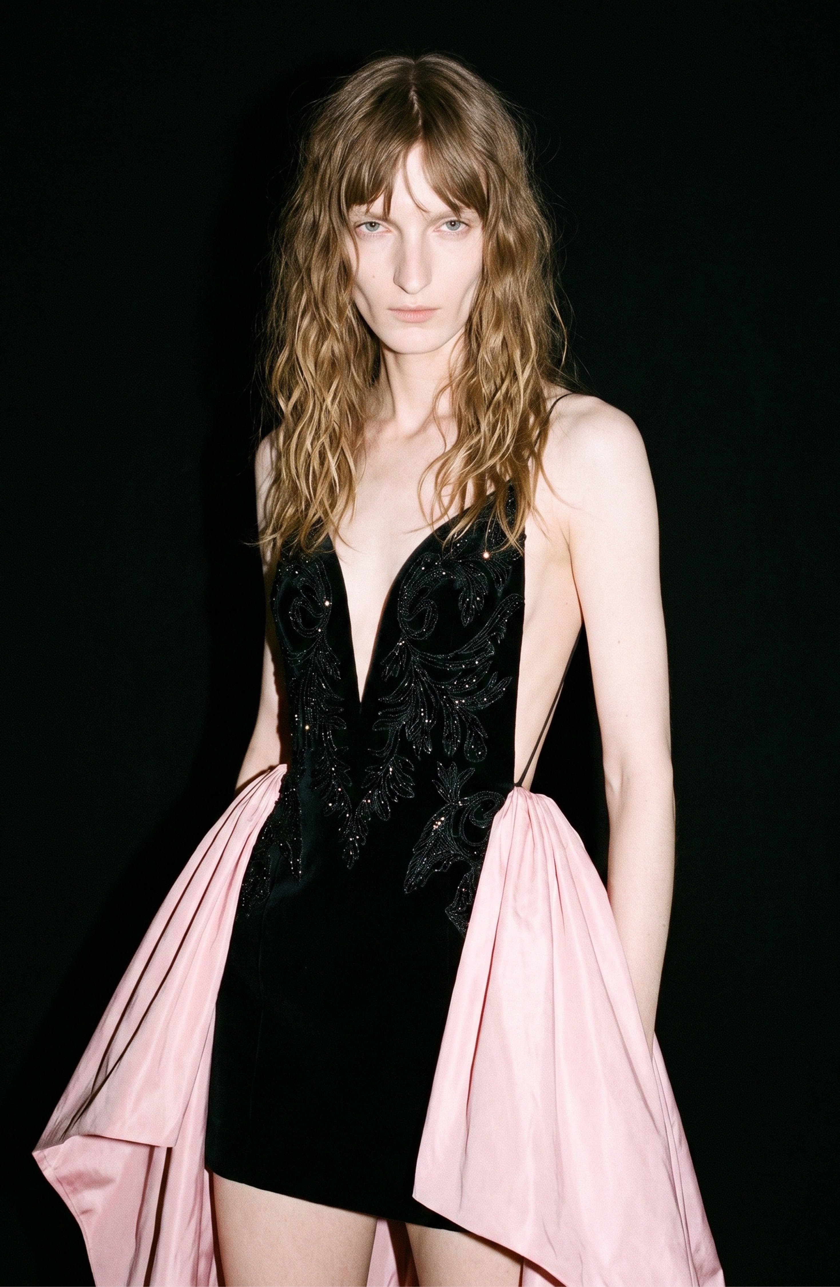 A person with wavy, light brown hair wears the LOULOU Embellished Velvet Mini Dress—featuring a deep V-neck, intricate detailing, and a flowing pink taffeta train—posing against a black background.
