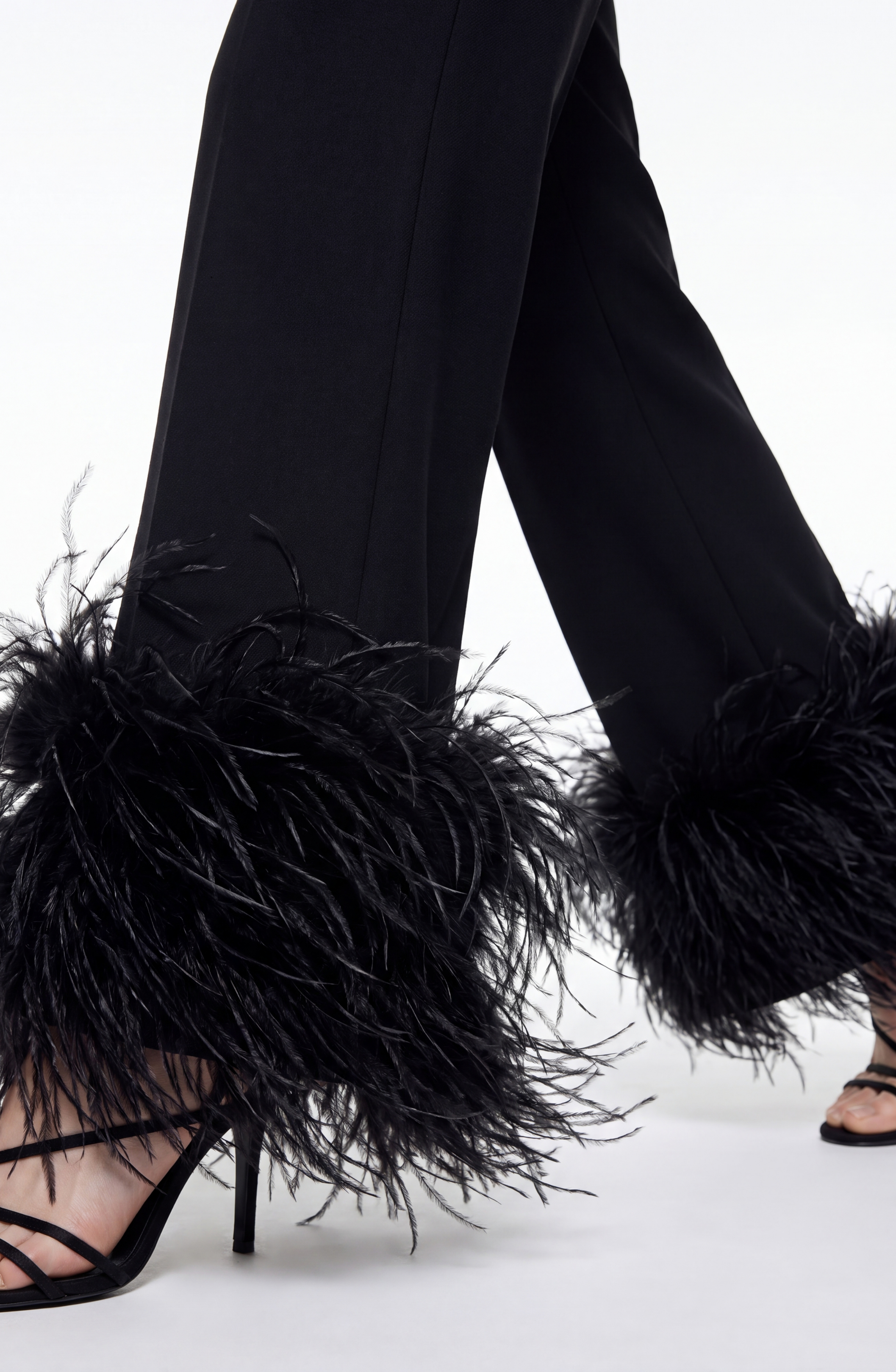 A person wears LOULOU Feather Trim Tailored Trousers featuring black feather detailing and straight-leg cuffs, paired with black high-heeled sandals, shown from mid-thigh down on a white background.
