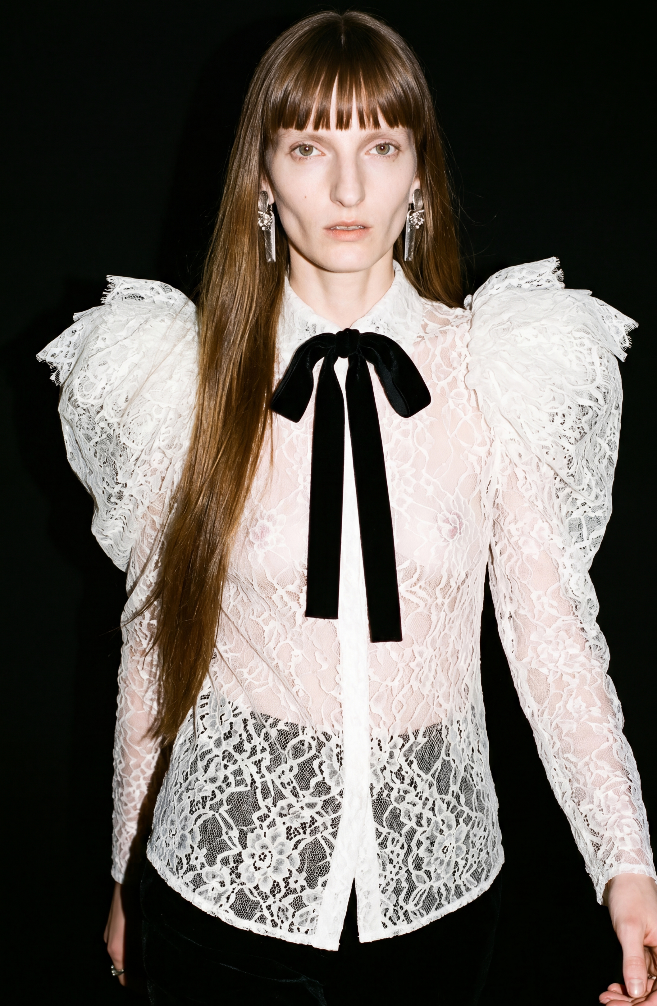 A person with long brown hair wears the LOULOU Lace Puffed Sleeve Blouse, featuring sheer French lace, dramatic puffed shoulders, and a black velvet bow, paired with statement earrings and set against a black background.