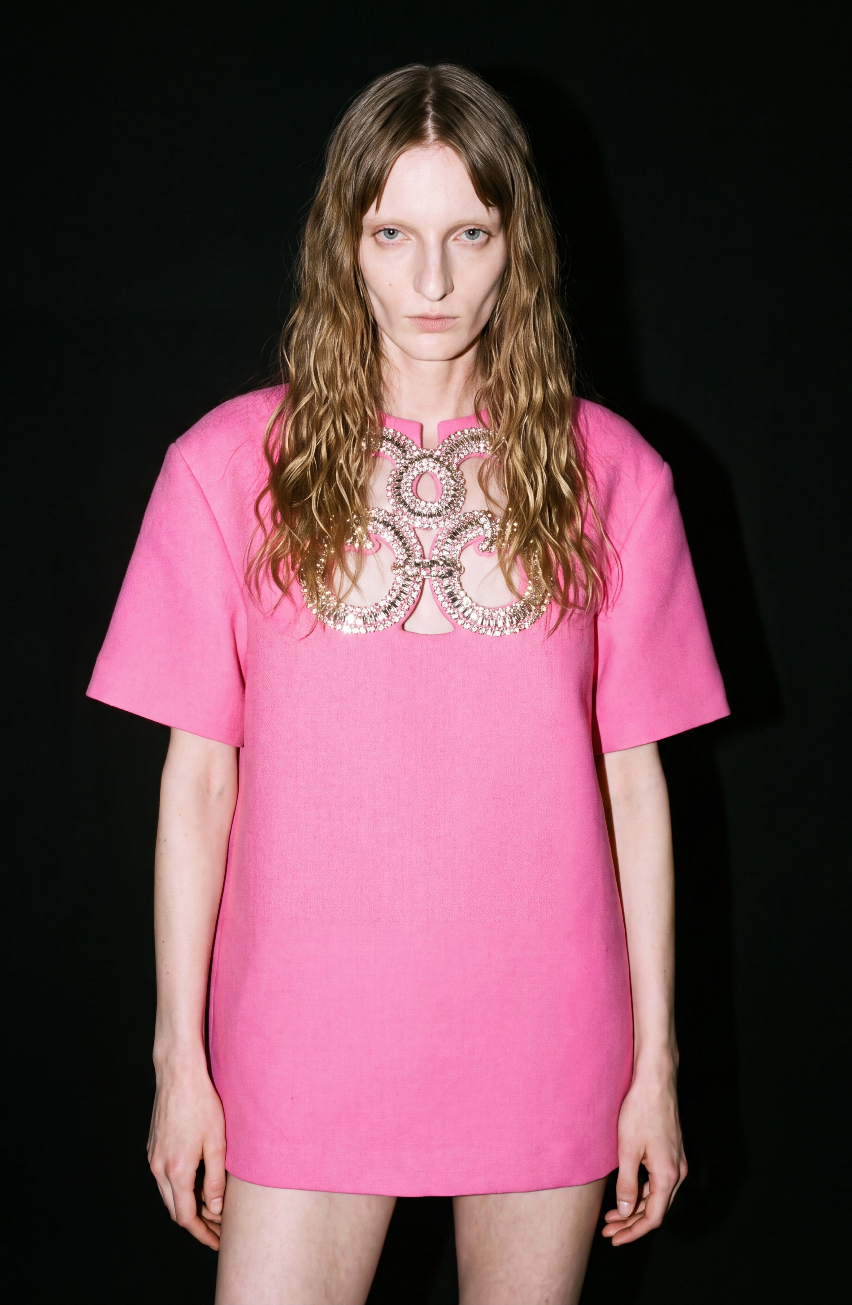 A person with long wavy hair wears the LOULOU Crystal Cutout Mini Dress, featuring crystal embellishments on the chest, as they stand against a solid black background.