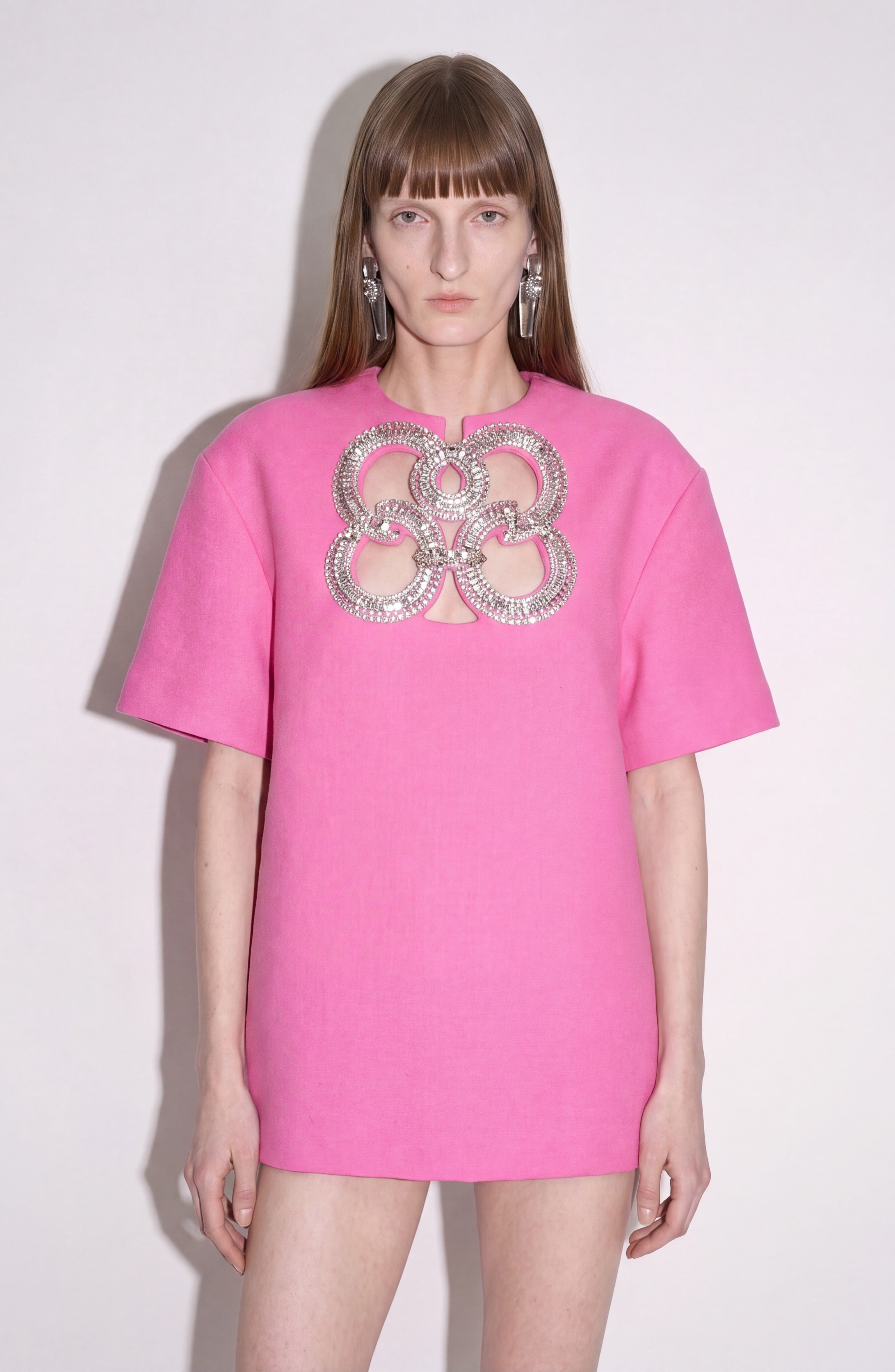A person wears the LOULOU Crystal Cutout Mini Dress, featuring a fuchsia hue, large circular silver embellishments, and a keyhole cutout on the chest, set against a plain light background.