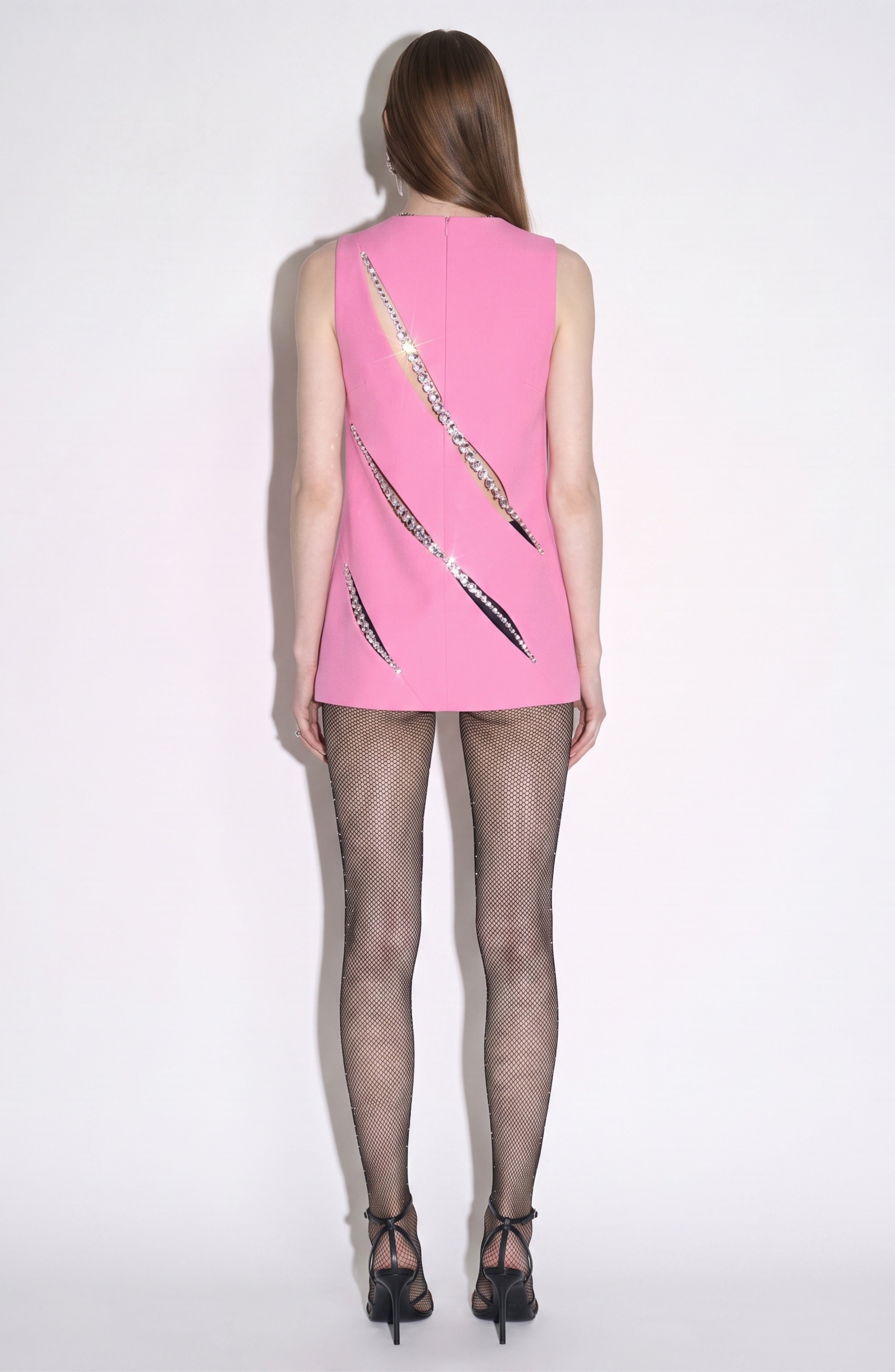 A person is seen from behind, wearing the LOULOU Peony Crystal Slash Top—a sleeveless pink tunic featuring striking shiny silver slashes on the back and a structured crepe design that adds to its sleek appearance. The look is finished with black fishnet tights and black high heels, set against a simple white background.