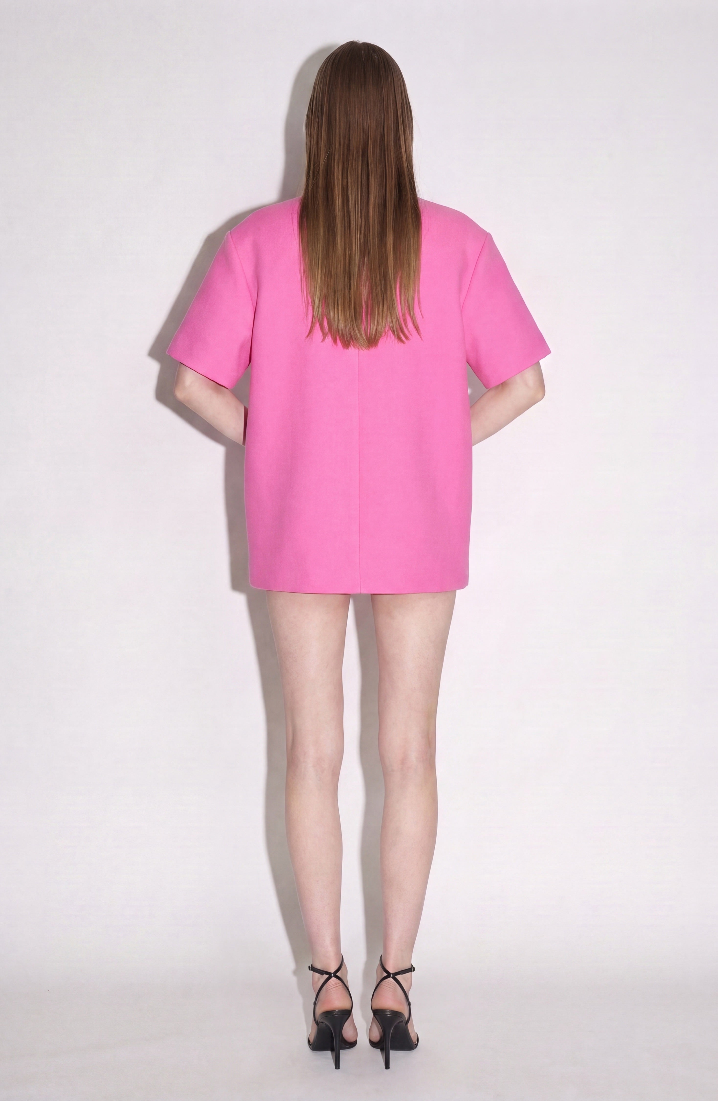 A person with long straight hair stands facing a white wall, wearing the LOULOU Crystal Cutout Mini Dress in vibrant fuchsia and black high-heeled sandals. Their arms are relaxed at their sides.