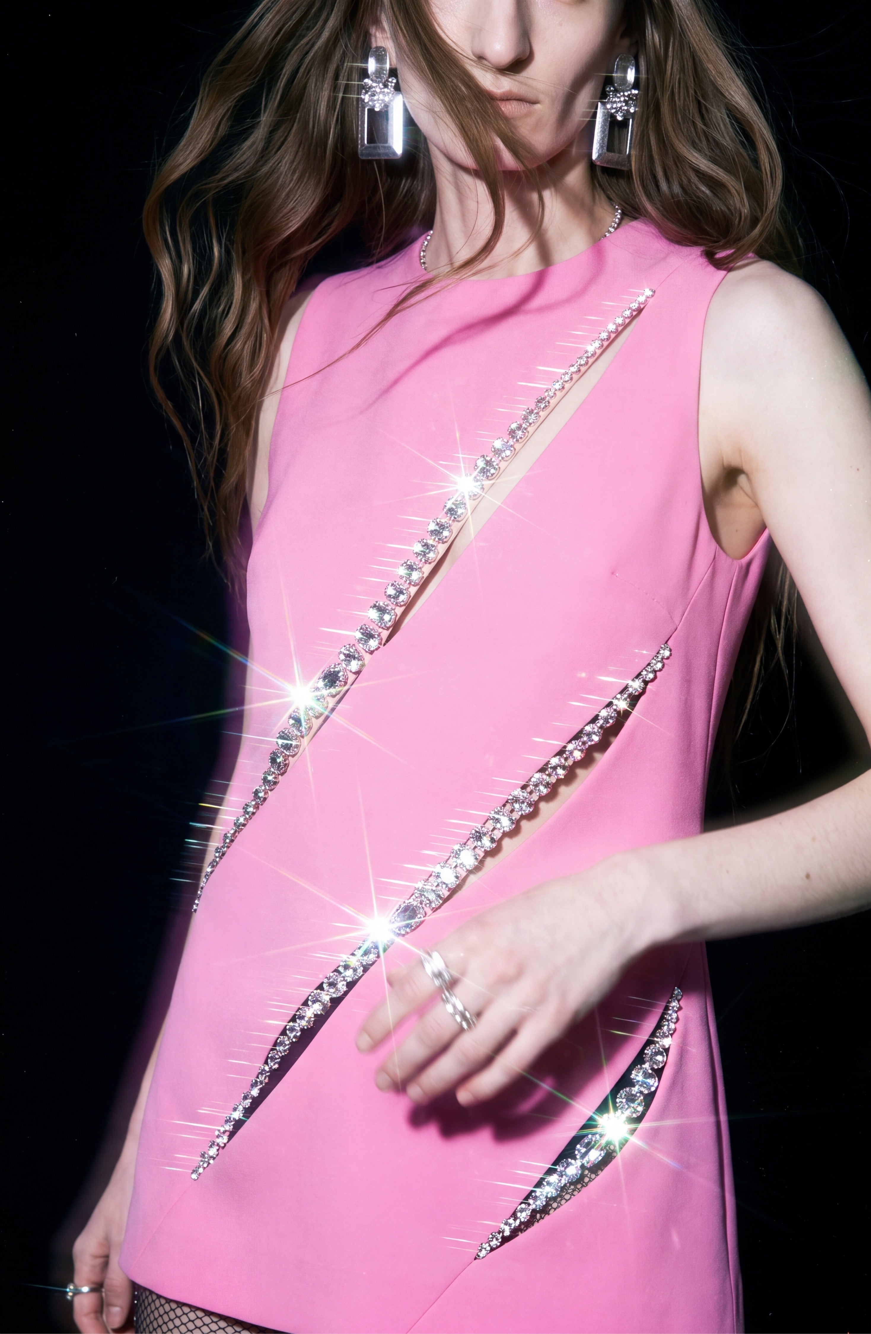A person wears the LOULOU Peony Crystal Slash Top—a sleeveless, shift silhouette pink top featuring diagonal rhinestone slashes—paired with large silver earrings and rings. The glass crystal trim glitters under bright light against a black background.